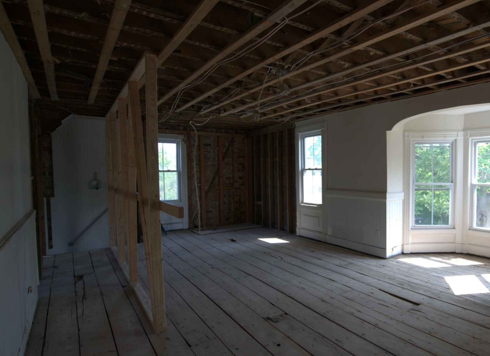 Interior of a room under renovation with exposed wooden ceiling beams, framing, and missing drywall. Natural light from multiple windows illuminates the space, which has a hardwood floor and partially finished walls. 