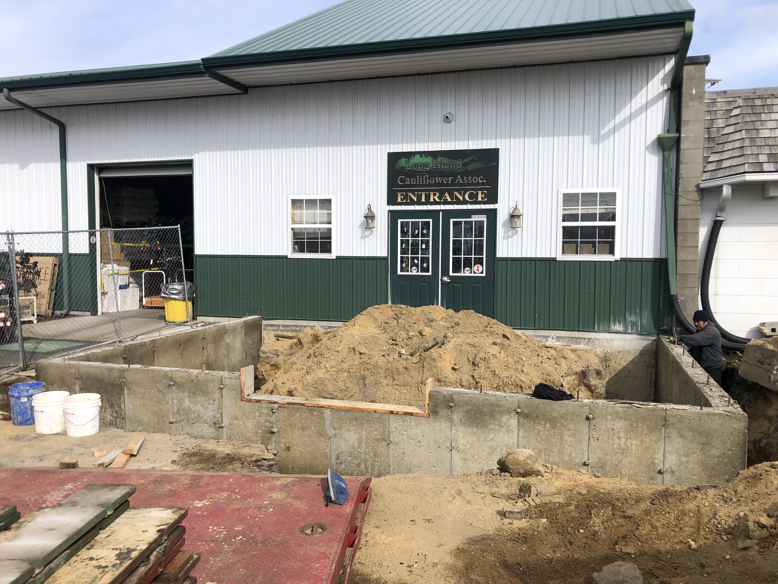 Ratsey Construction site in front of the Long Island Cauliflower Association building, with dirt piles, a concrete foundation, and a worker on the right.