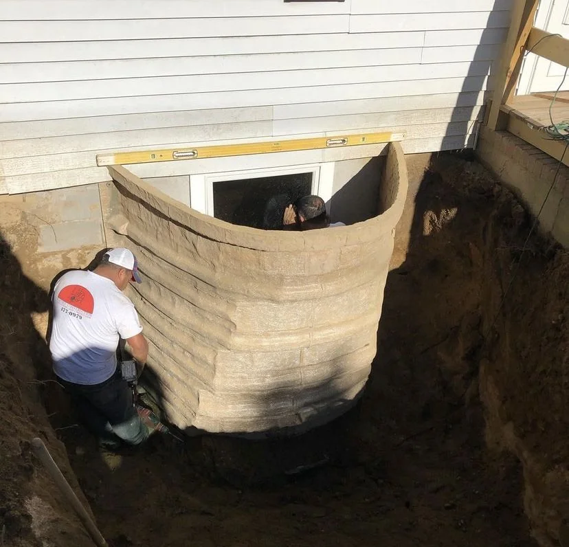 Construction workers installing a curved stone wall at the foundation of a house, with one worker inside a basement window and another outside supporting the structure.