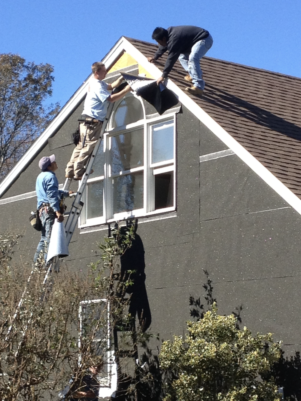 Three Ratsey workers installing or repairing an arched window on the upper floor of a house with a steep roof and shingle siding.
