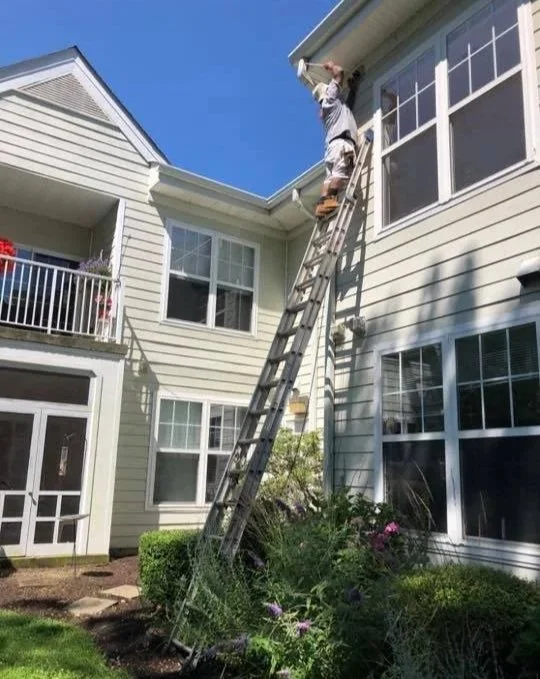 A person standing on a tall ladder leaning against the house, working near the roof of a multi-story residential building with beige siding and large windows.