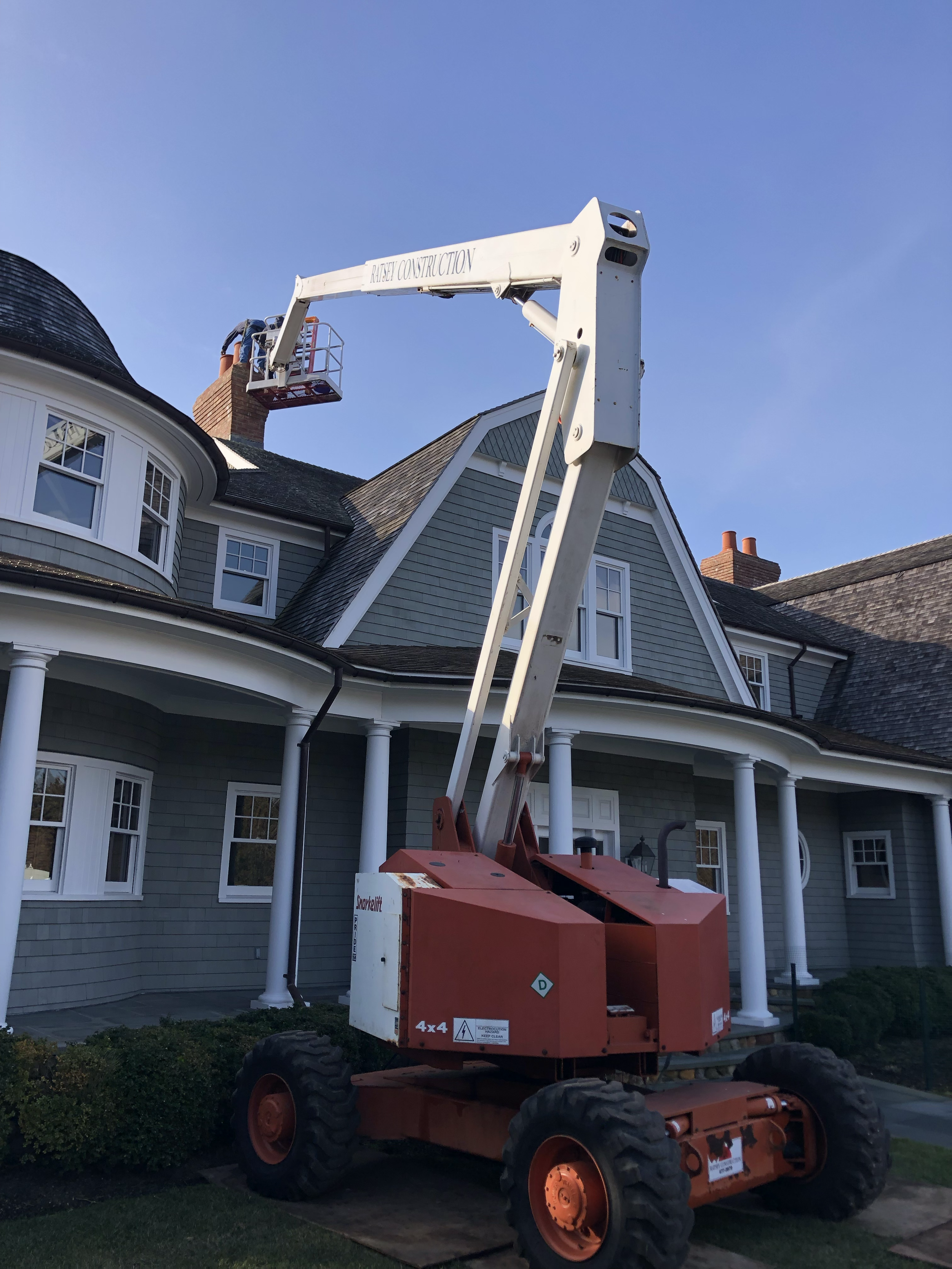 A cherry picker lifting a worker near the chimney of a large, multi-story house with grey siding and white trim.