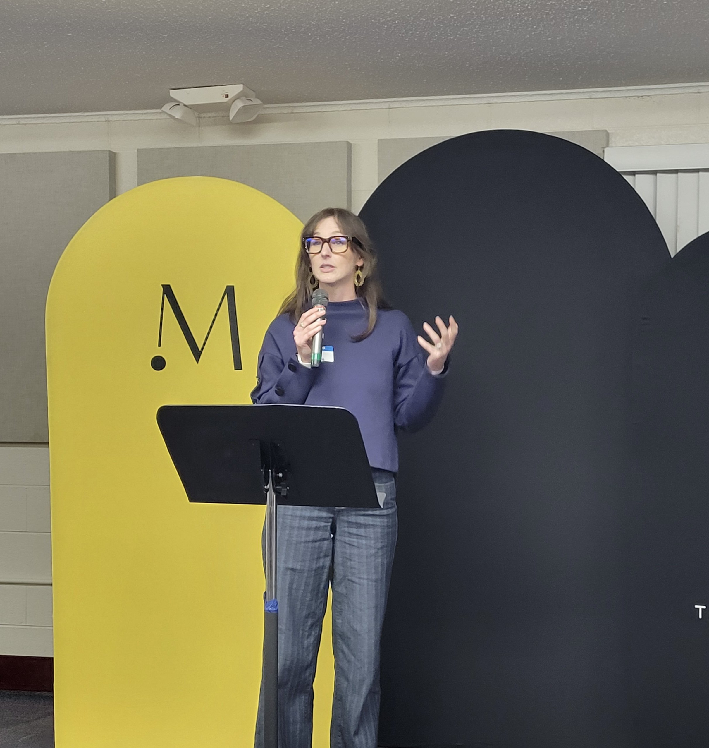 A woman with glasses, wearing a blue top and plaid pants, is speaking into a microphone while standing behind a black music stand. She is at a presentation event with large yellow and black decorative panels in the background.