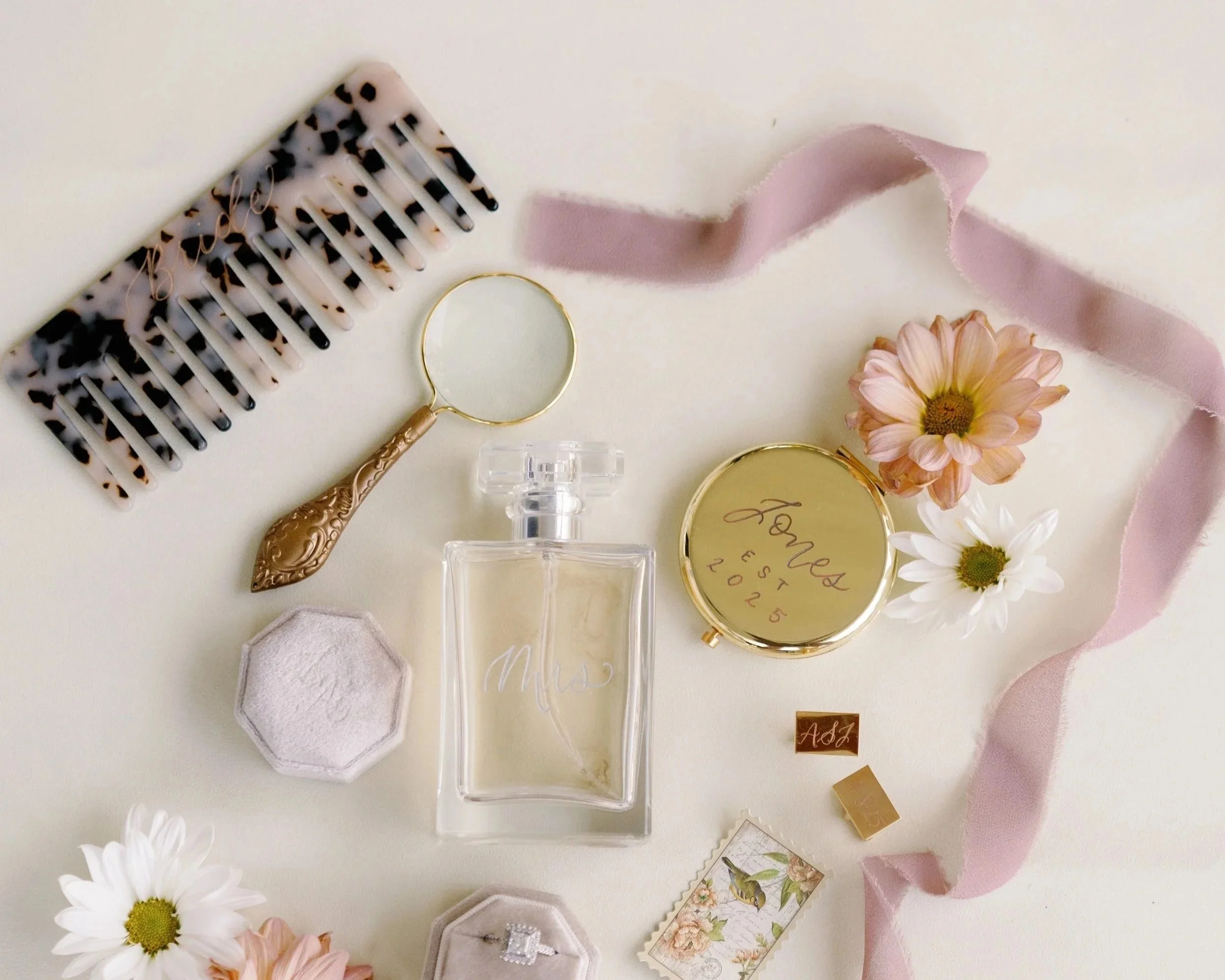 Flat lay of wedding accessories including a tortoiseshell hair comb, a round mirror, a perfume bottle labeled 'Mrs.', a pink flower, a white flower, a pink ribbon, a gold compact with 'James Est 2025' engraved, a small hexagonal container, a ring box