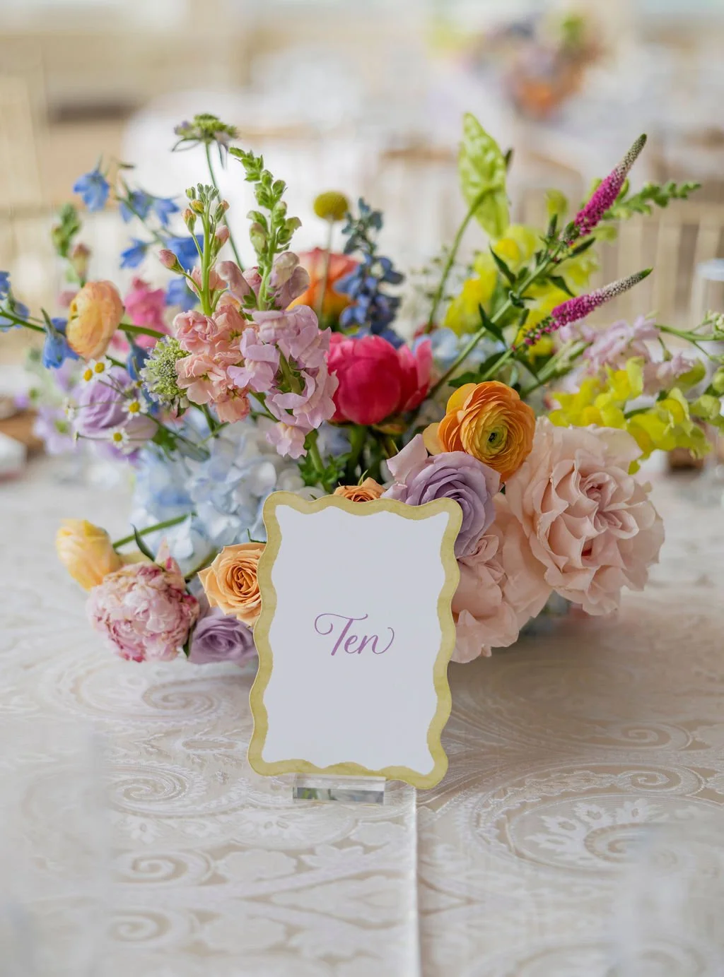 A colorful flower arrangement with pink, purple, yellow, and blue blooms on a table with a white tablecloth. A small, decorative sign reading 'Ten' is in front of the flowers, indicating the table number.