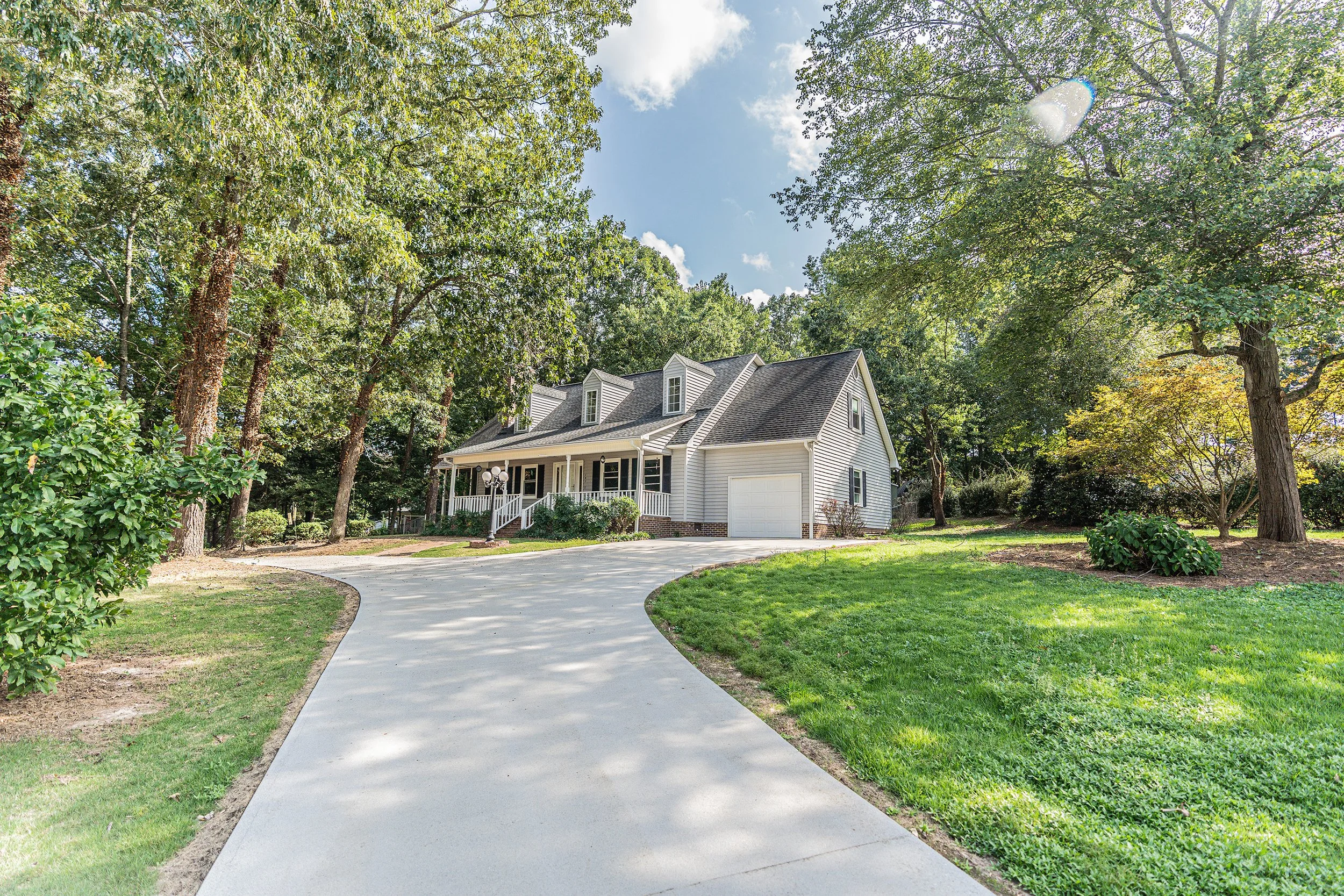 A house with a driveway in a lush green yard, surrounded by trees and a clear blue sky.