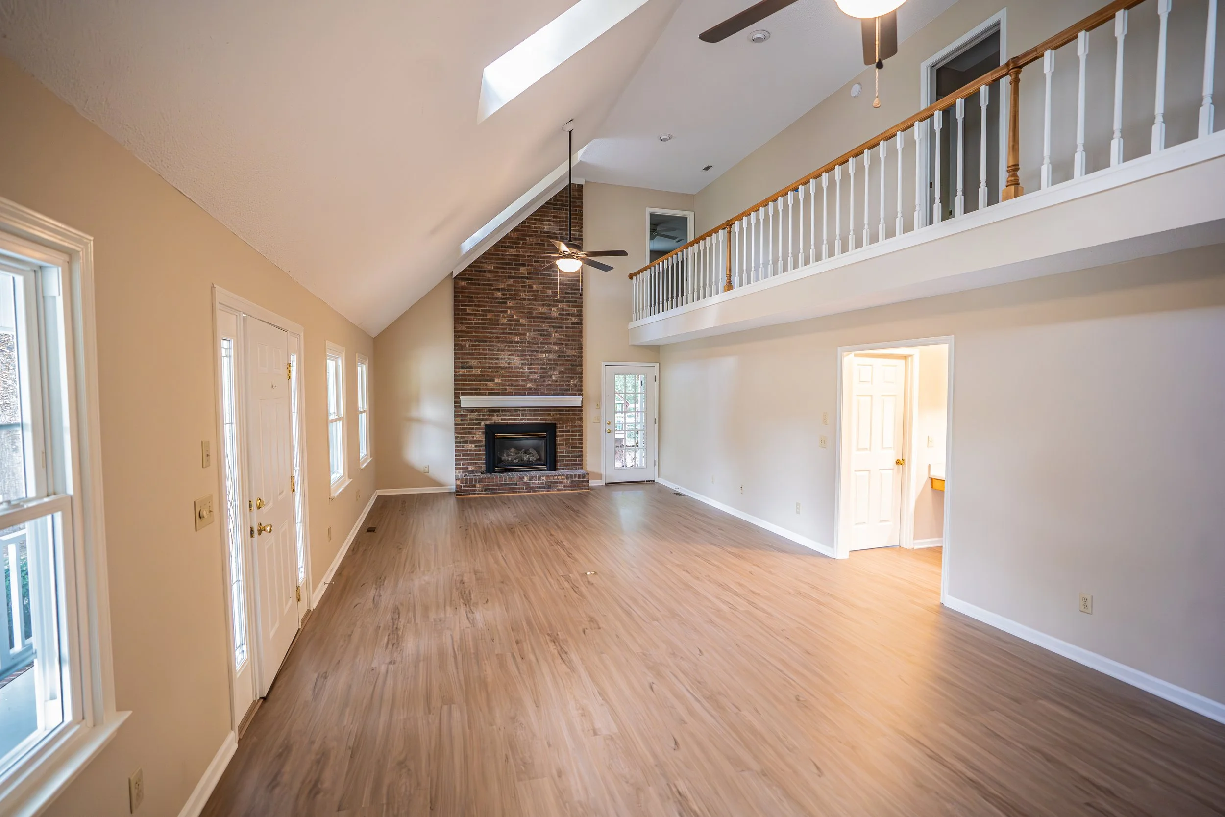 Empty living room with wooden floors, a brick fireplace, multiple windows, ceiling fan, upper balcony with railing, and a door leading outside.