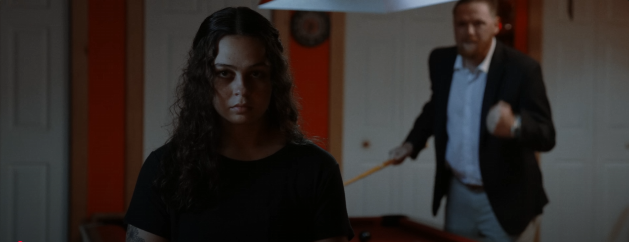 A young woman with dark, curly hair looking serious in the foreground, and a man in the background with a cue stick in hand, appearing to play pool in an indoor game room.