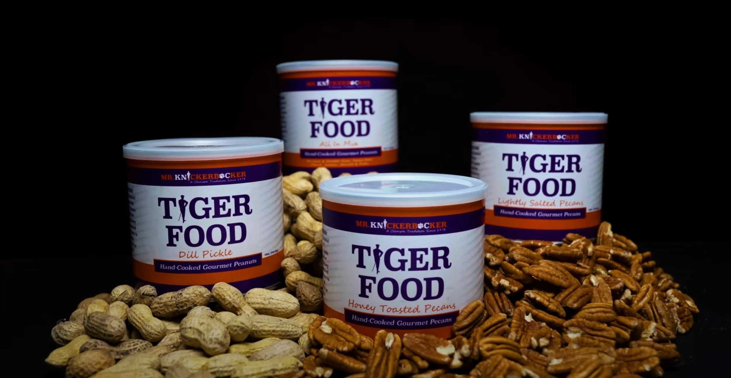 Four cans of Tiger Food brand gourmet pecans and peanuts arranged on a black background with peanuts and pecans spilled around them.