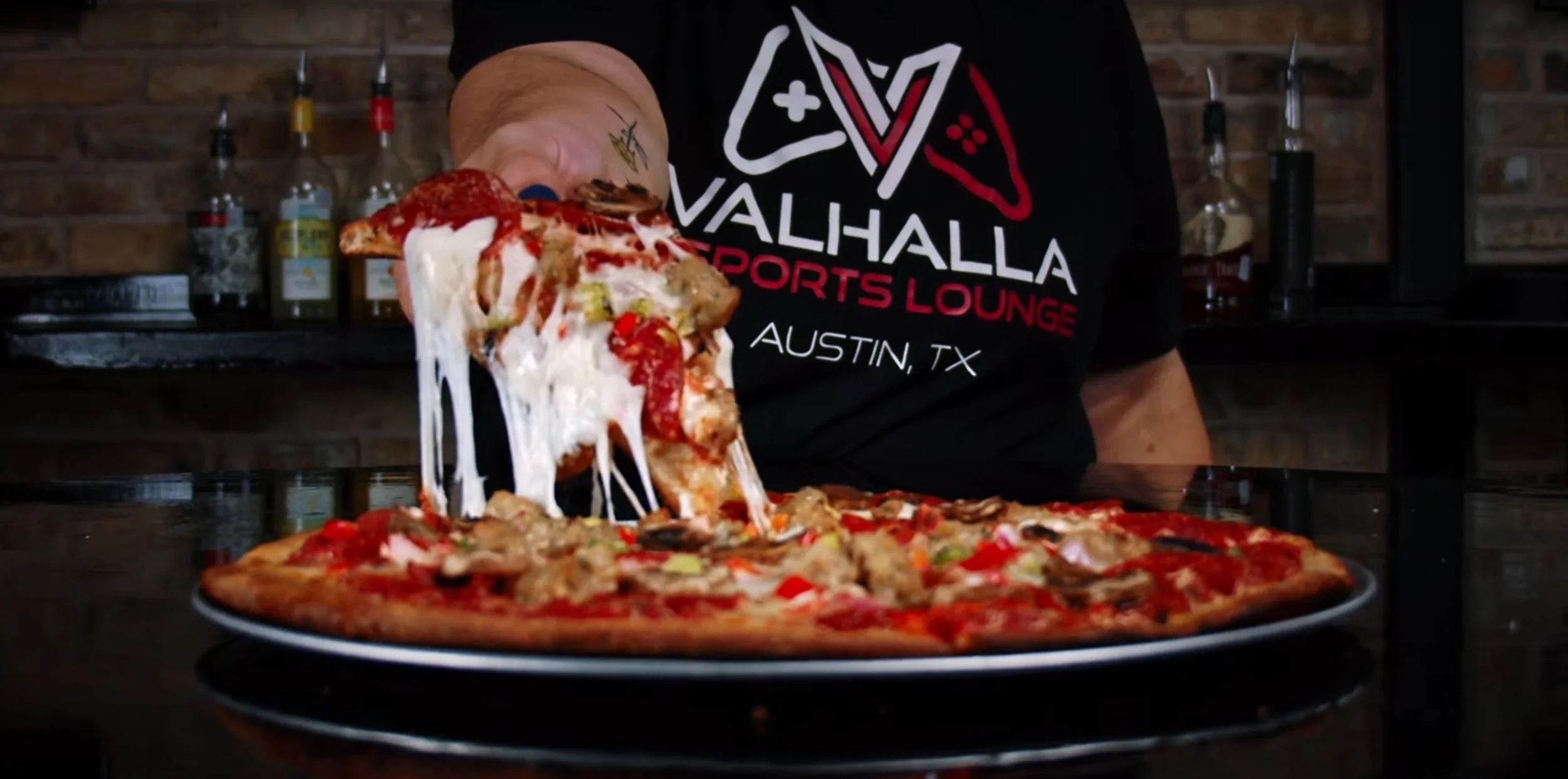Person holding a slice of pizza with melted cheese, pepperoni, vegetables, and mushrooms, with the rest of the pizza in front on a black table, in a restaurant setting.