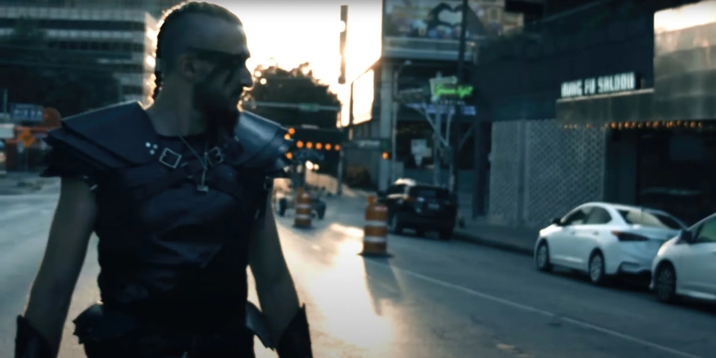 A person dressed in black futuristic armor with braided hair, walking on a city street during sunset, with cars parked and buildings in the background.