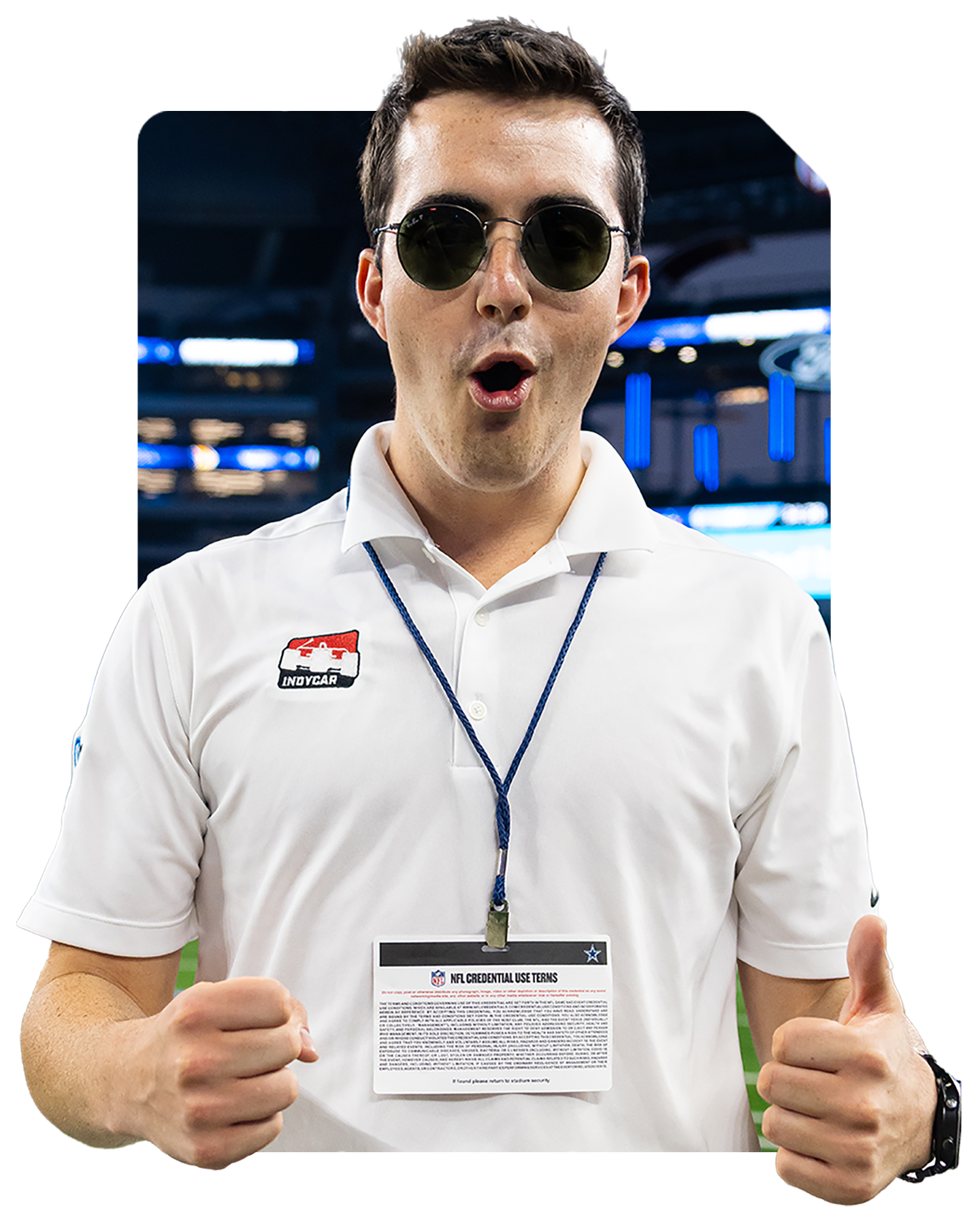 A man wearing sunglasses and a white polo shirt with an NFL credential lanyard, giving a thumbs-up gesture. He is indoors, possibly at a sports event or arena.