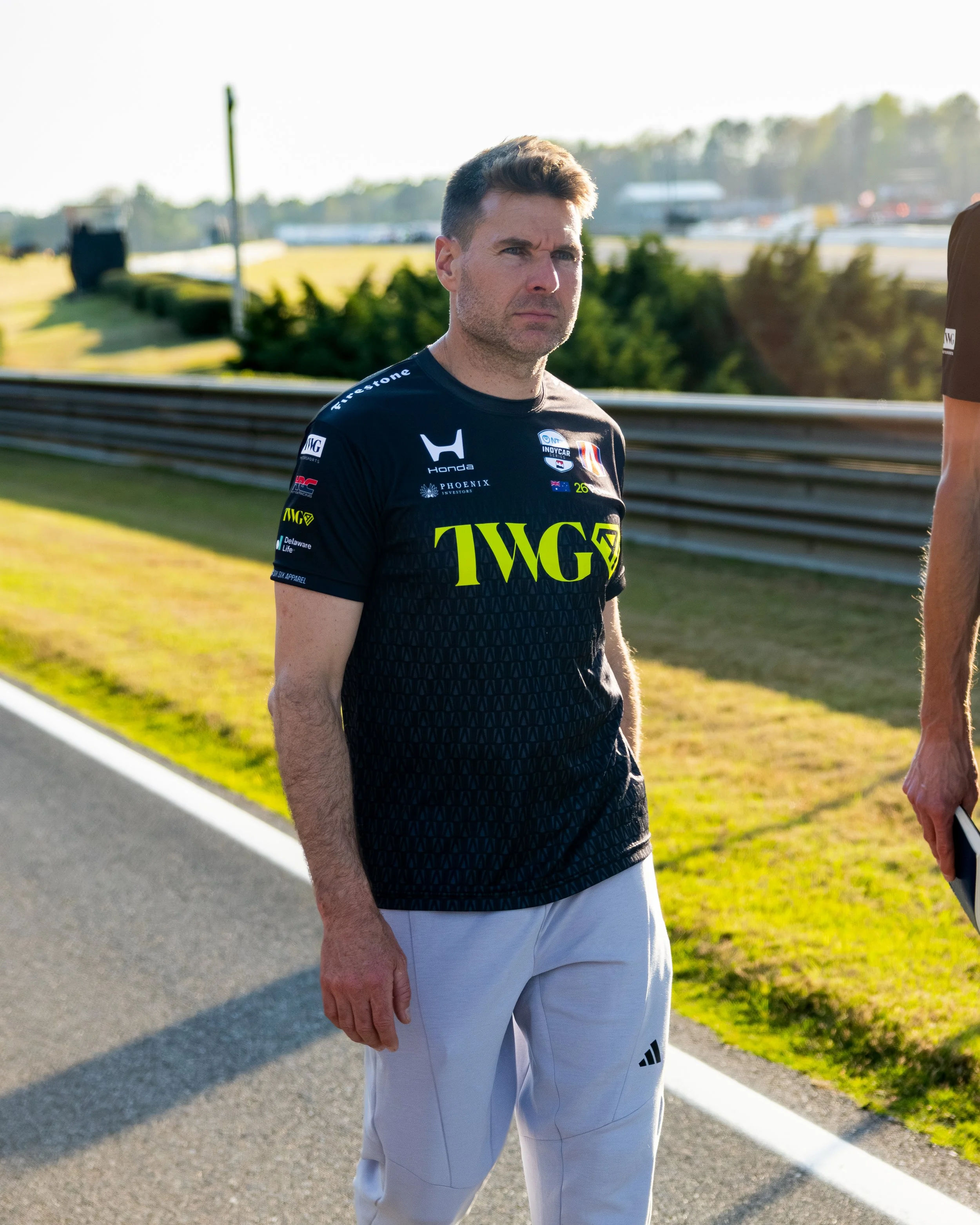 A man with short brown hair and a serious expression standing outdoors near a racetrack with a safety barrier, wearing a black racing shirt with various sponsor logos and white pants, with sunlight casting shadows.