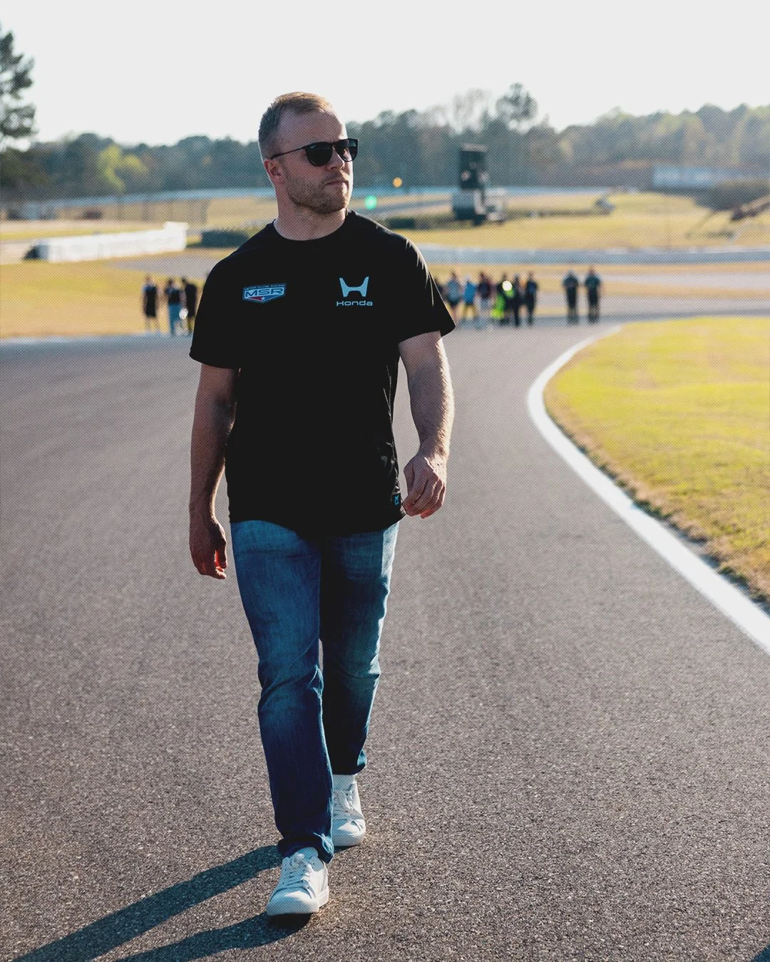 Man walking on racetrack in daytime, wearing sunglasses, black t-shirt with Honda and MSR logos, jeans, and white sneakers, with group of people in the background.