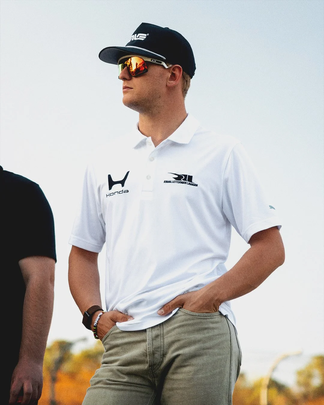A man wearing a white polo shirt, black cap, and reflective sunglasses, standing outdoors with his right hand in his pocket. The shirt has logos including Honda and Rahal Letterman Lanigan and features short sleeves. The background shows a partly clo