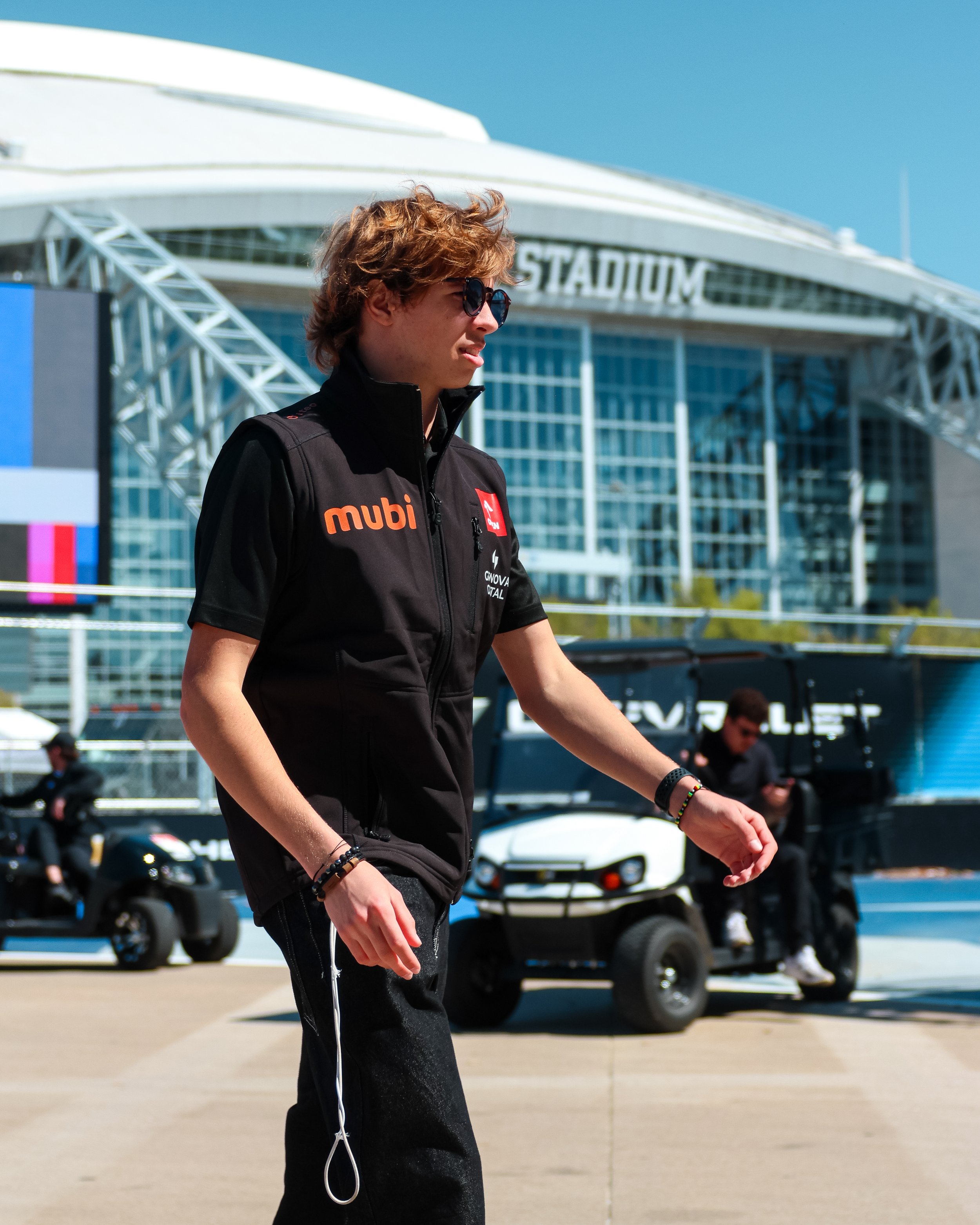 Young man with curly hair and sunglasses walking outdoors near a modern stadium, wearing a black jacket with sponsor logos.