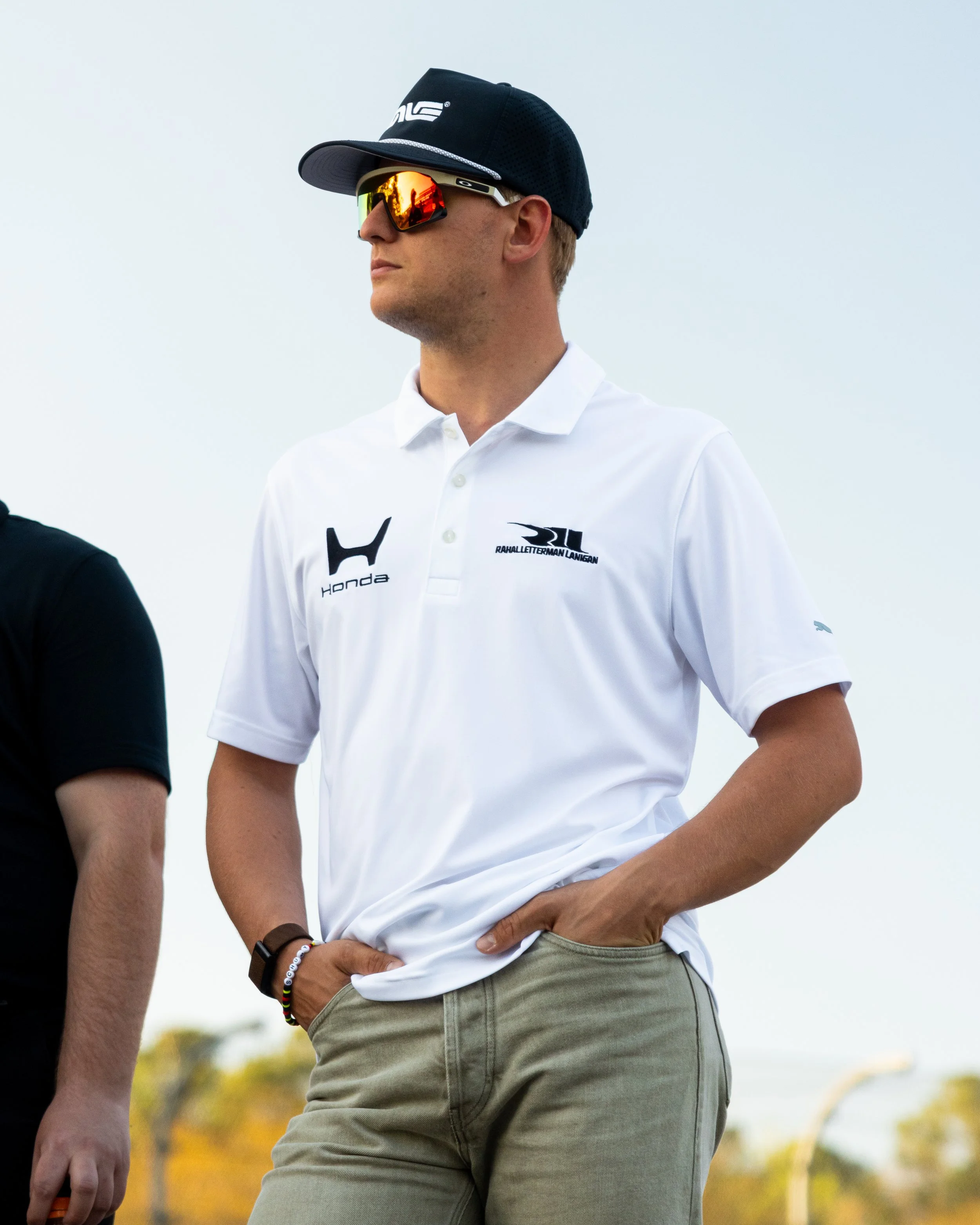 A person wearing sunglasses, a black cap, a white polo shirt with logos, and khaki pants standing outdoors during daytime.