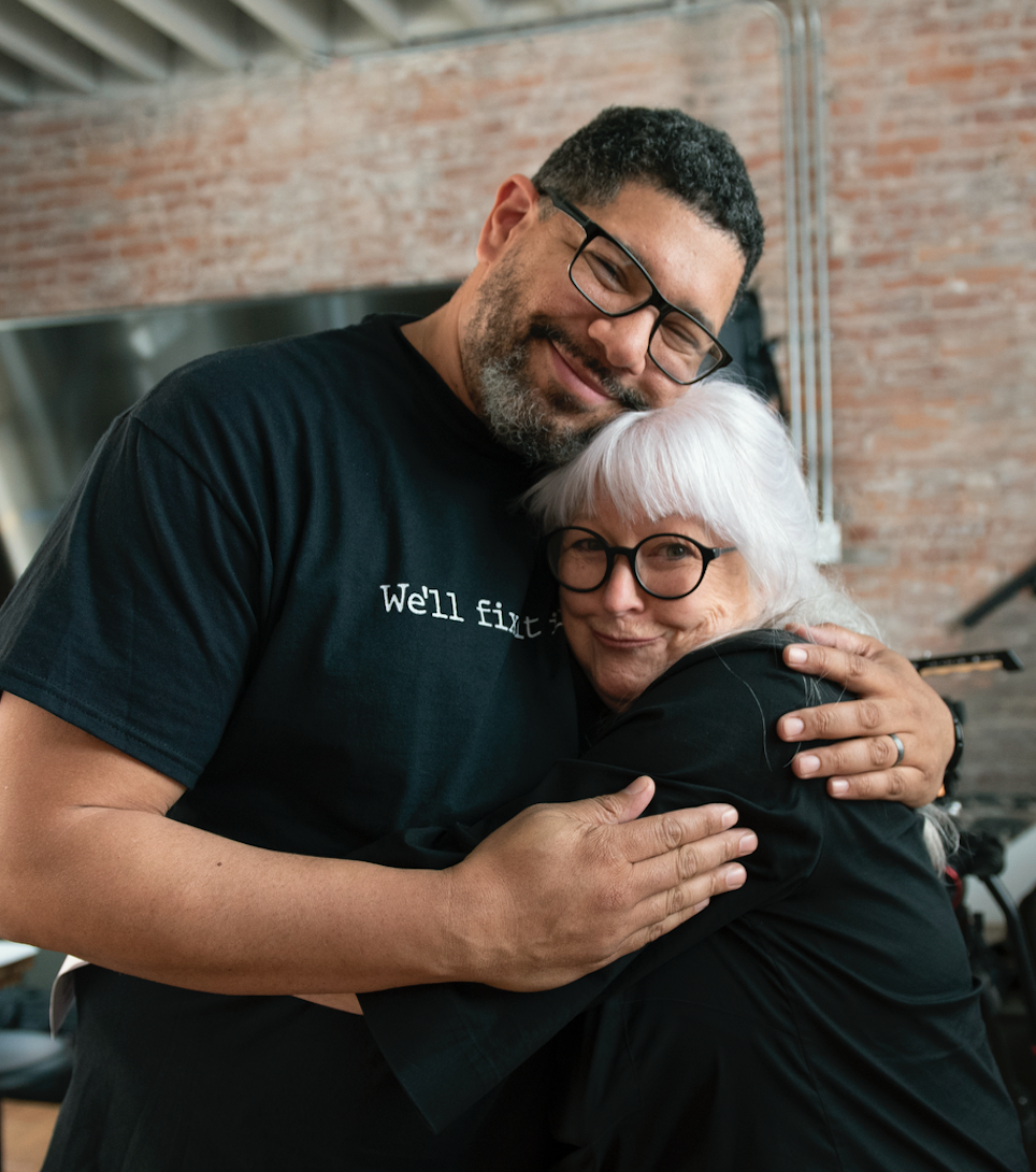 A man and an elderly woman hugging each other happily indoors with brick walls in the background.