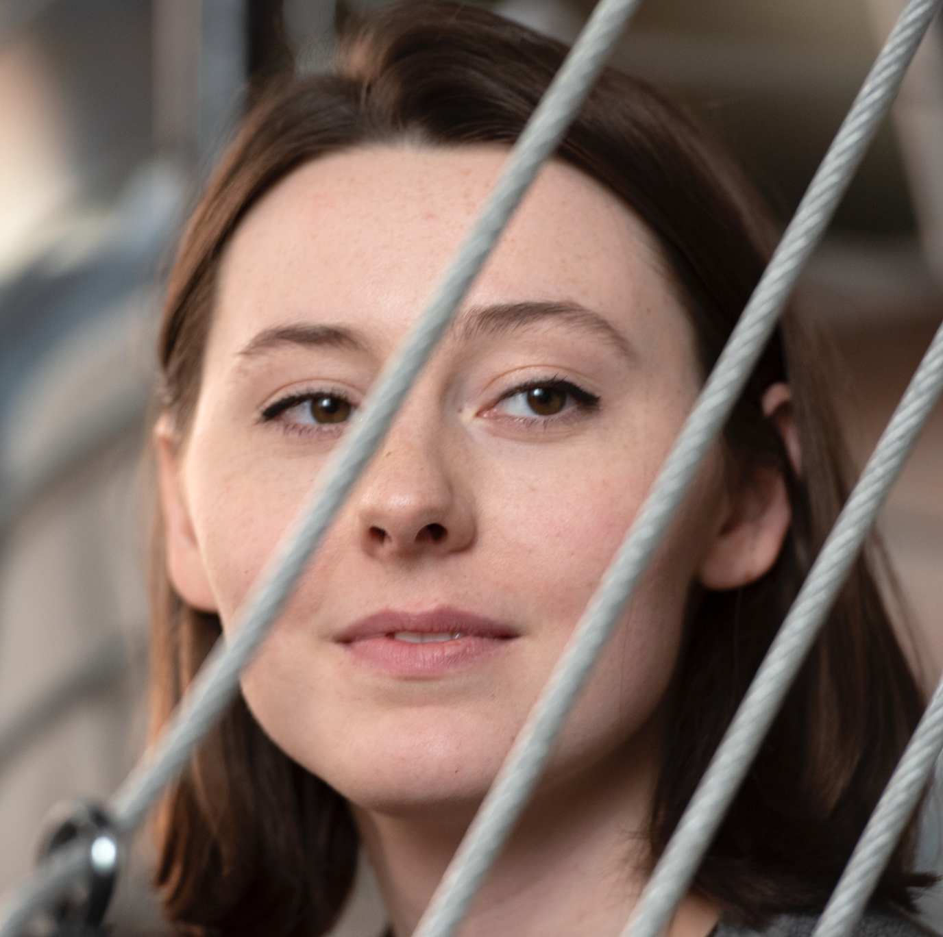 Close-up of a woman with brown hair and brown eyes, partially obscured by metal bars or wires.