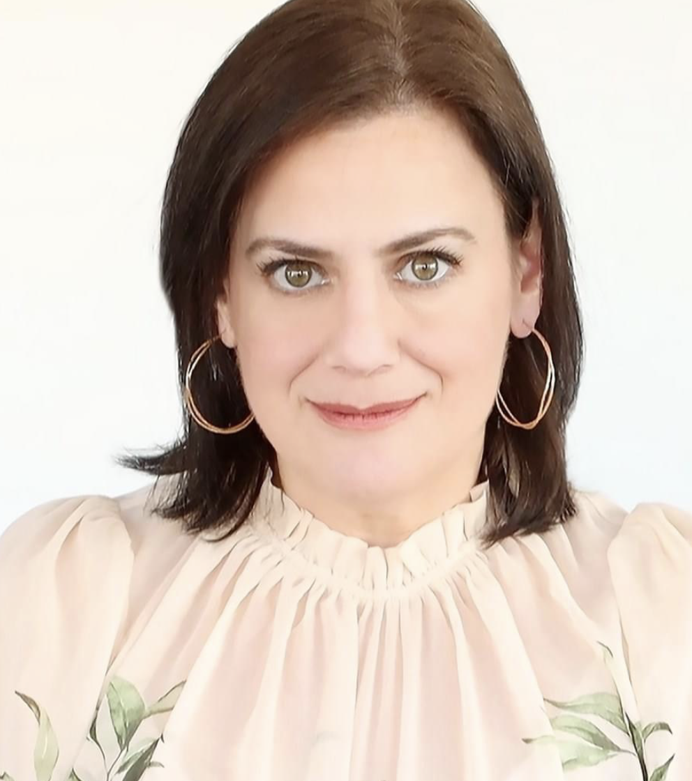 A woman with shoulder-length brown hair, gray-green eyes, and hoop earrings, wearing a beige blouse with a ruffled neckline, smiling at the camera against a plain white background.