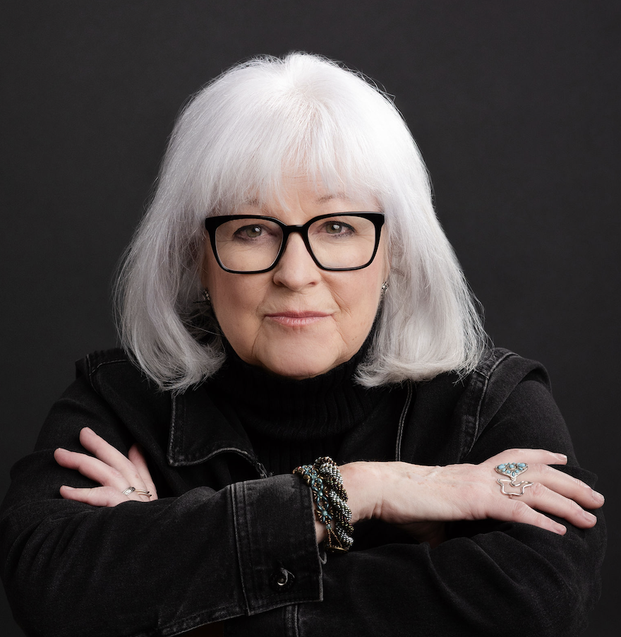 Johanna Beale Keller, award-winning playwright, filmmaker & journalist, who is a woman with shoulder-length white hair and black glasses wearing a black jacket, with her arms crossed, against a dark background.