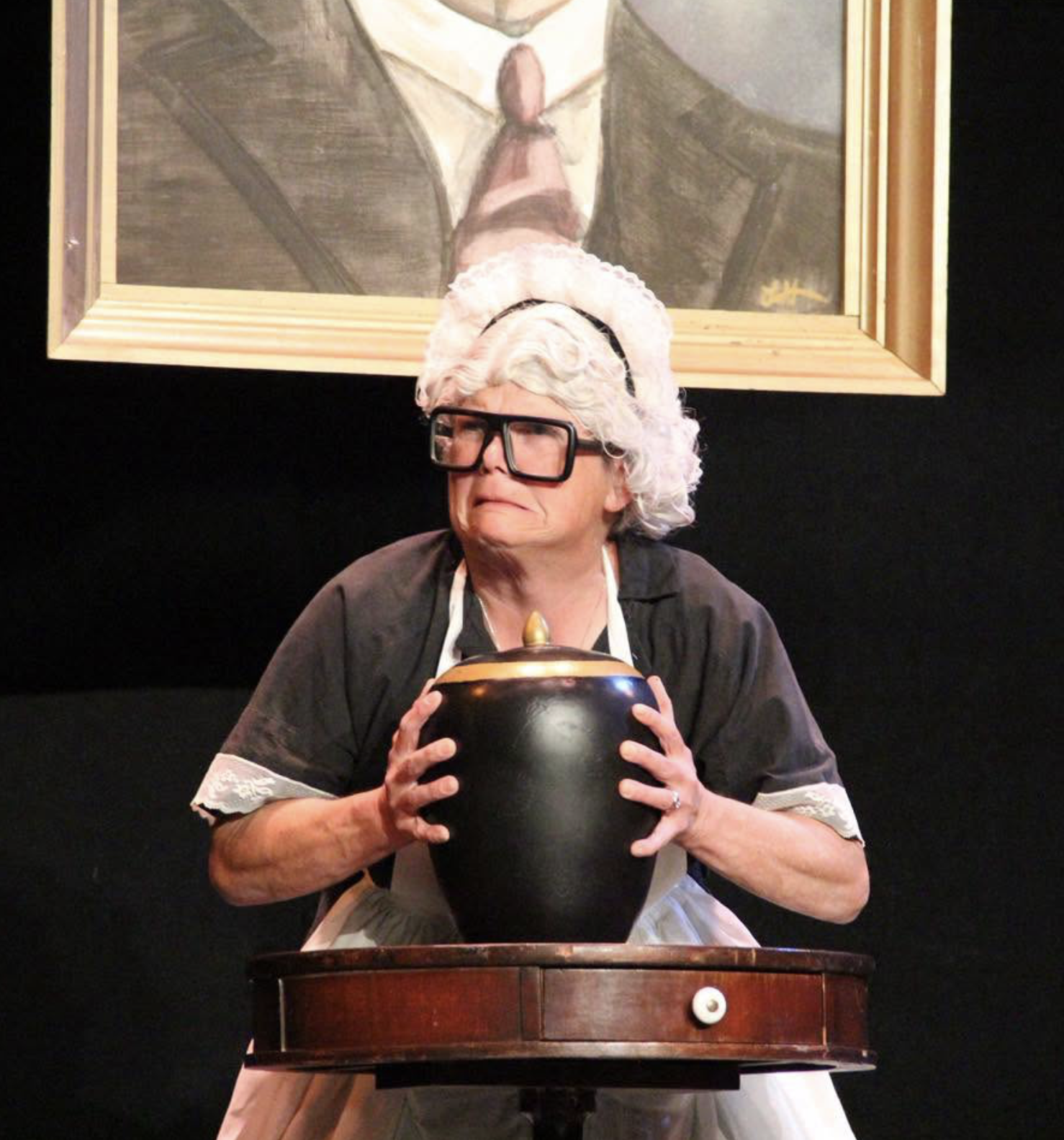 A person dressed as an elderly woman with white curly hair, large glasses, and a black dress, holding a black cauldron with both hands. They are sitting at a wooden table in front of a dark background, with a framed portrait of a man in a suit hanging on the wall behind them.
