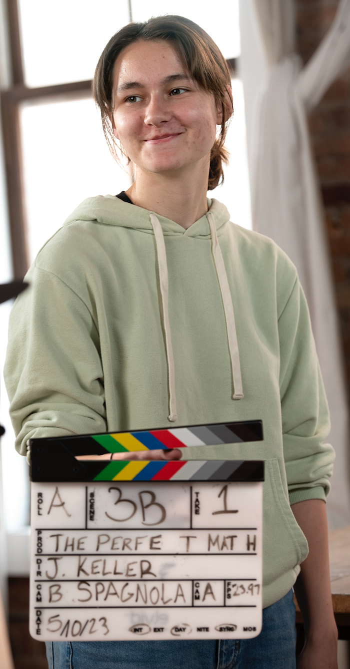 Young woman holding a film slate, smiling, in a bright room with large windows and brick wall in the background.