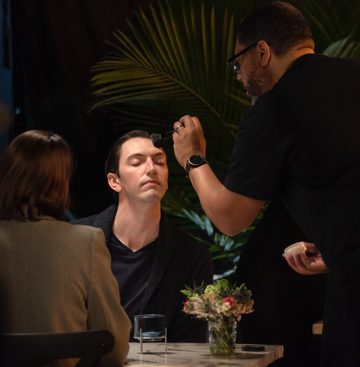 A man is having makeup applied to his face by a makeup artist at a table with a flower arrangement, glass of water, and a small container.