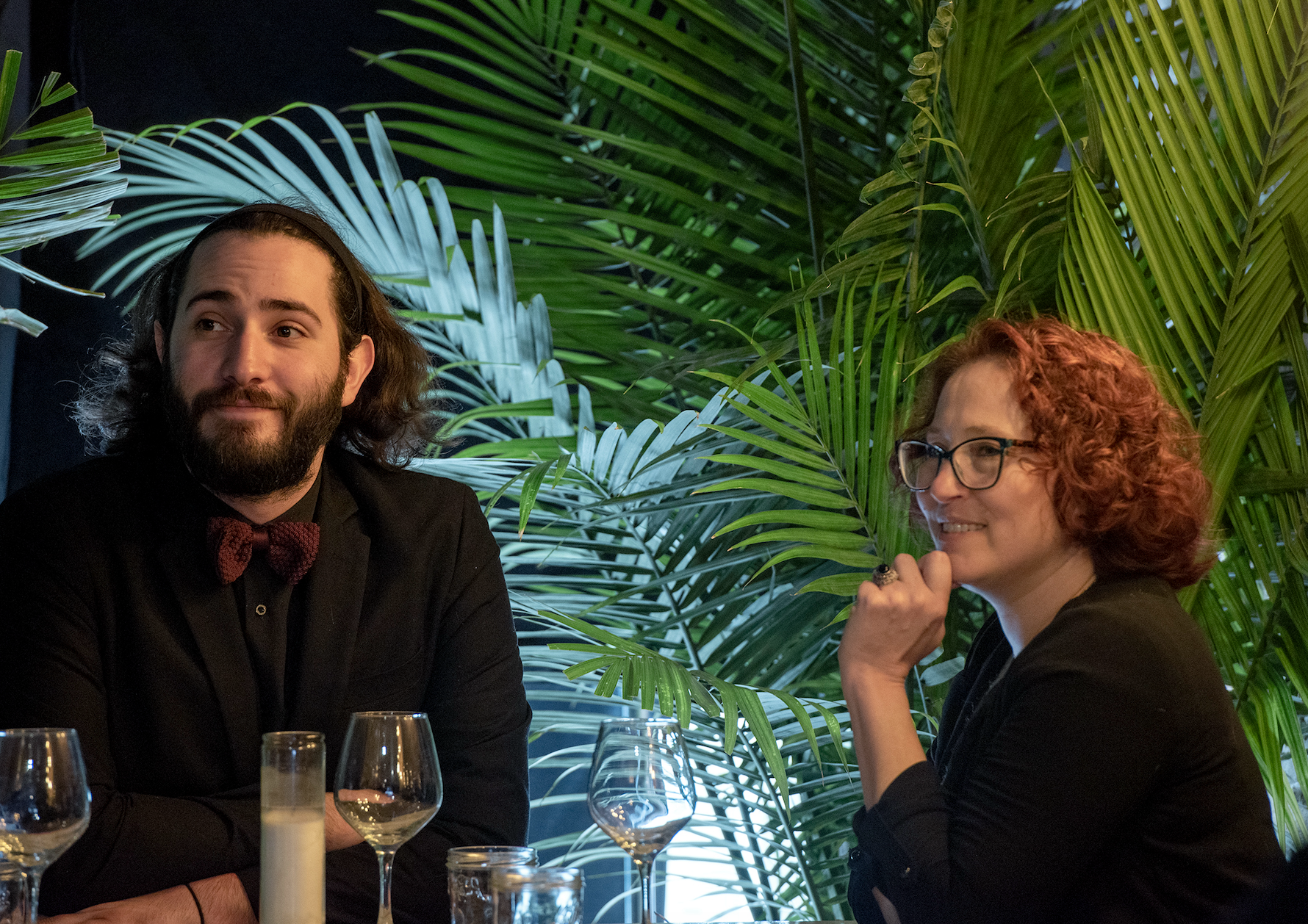 A man with long hair and a beard, wearing a black shirt and a red bow tie, sitting at a table with wine glasses and candles, smiling and looking to his left. Next to him, a woman with curly red hair, glasses, wearing a black jacket, resting her chin on her hand, smiling, with large green palm leaves in the background.