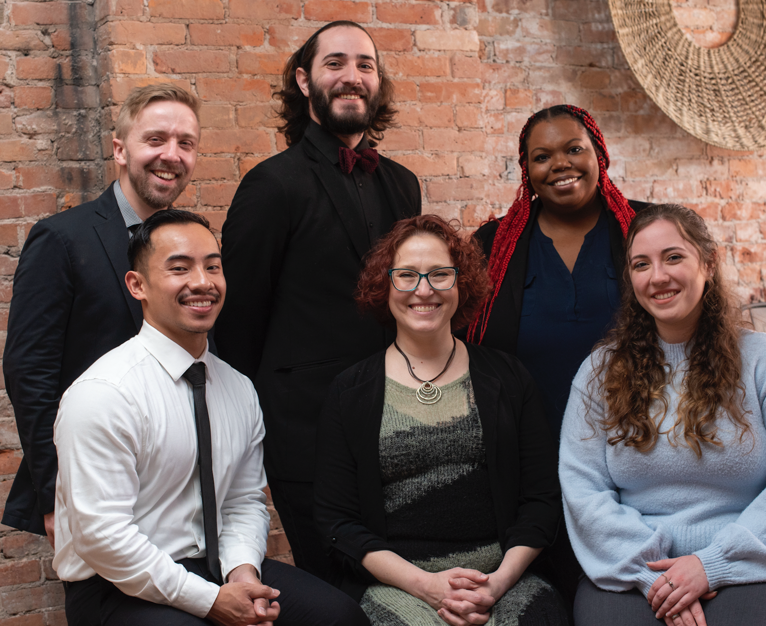 Group portrait of seven diverse young professionals indoors against an exposed brick wall, smiling and dressed in business casual attire.