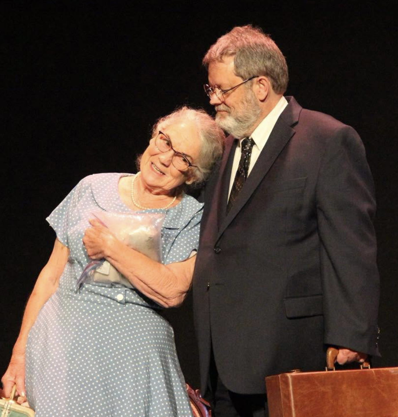An elderly woman with glasses and a pearl necklace, wearing a blue polka-dot dress, smiling and holding a bouquet of flowers, stands next to a man in a dark suit and tie, who is holding a brown handbag, both on a stage with a black background.