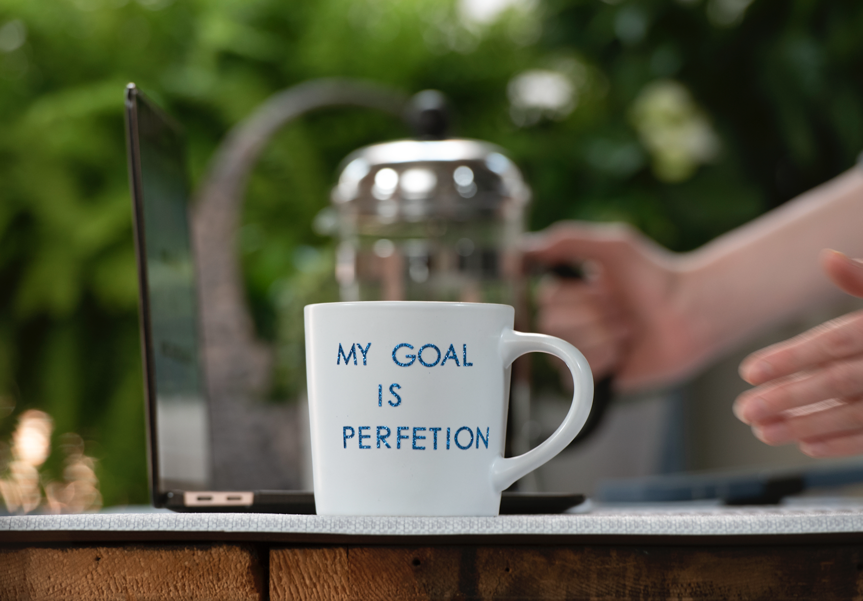 A white coffee mug with the text 'MY GOAL IS PERFECTION' on it, placed on a table with a laptop and a person typing in the background. The setting appears to be outdoors or in a garden with greenery.