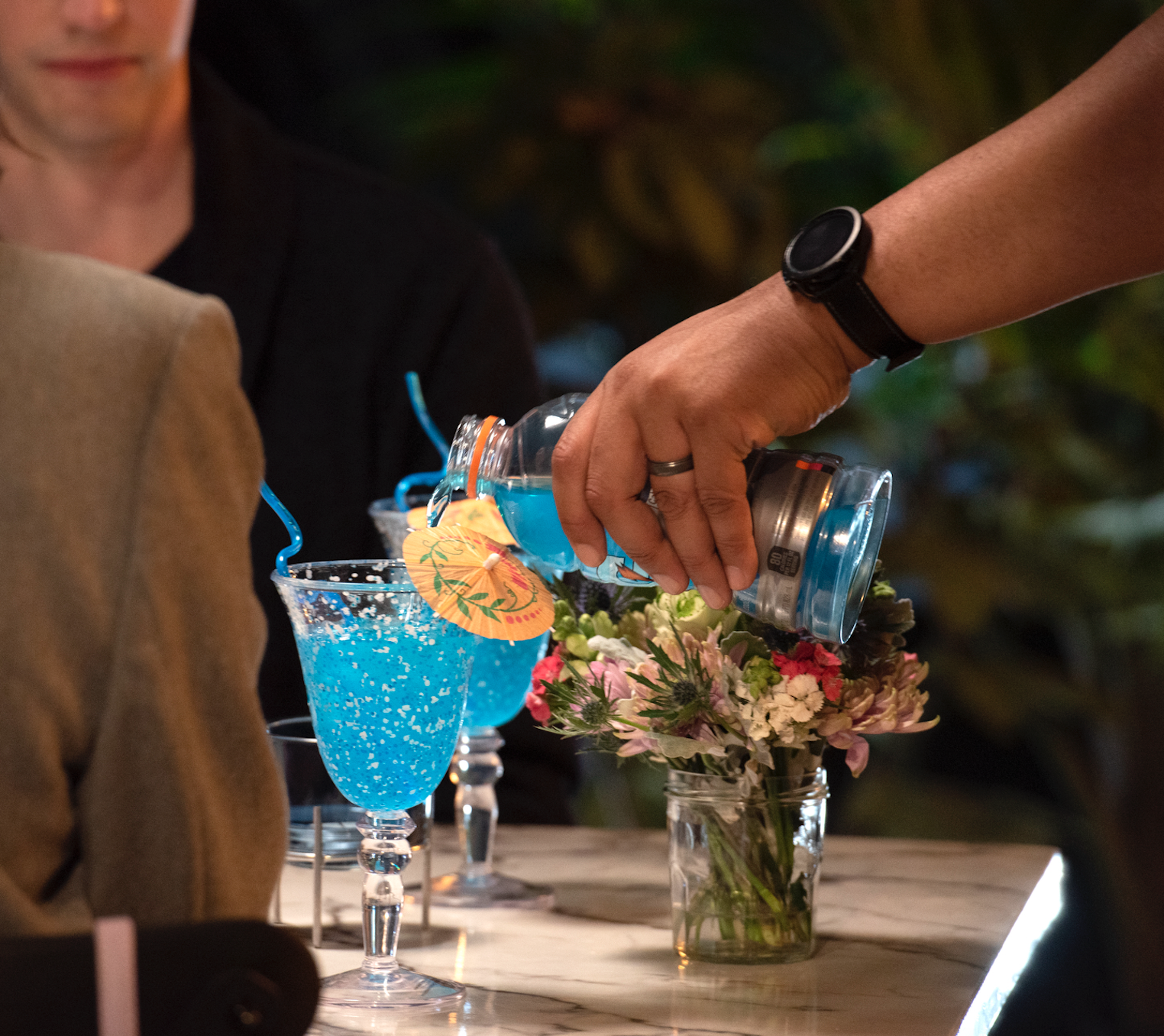 Person pouring a blue cocktail into a glass with an umbrella at a table with floral arrangements