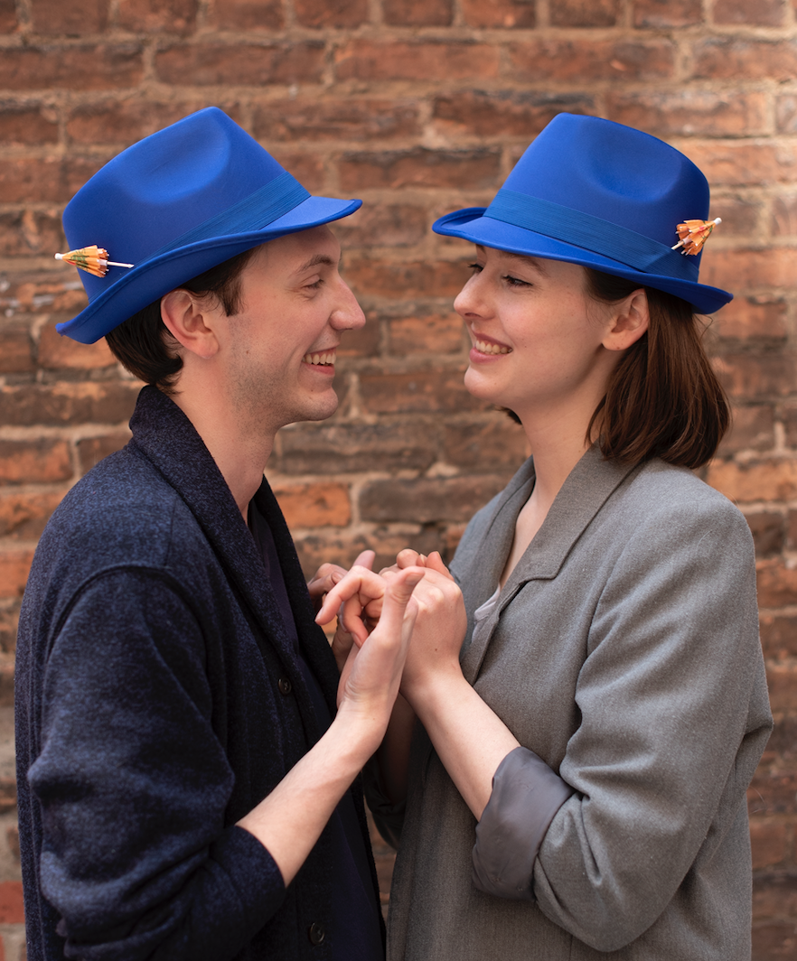 A man and woman wearing blue hats with small umbrellas attached, smiling and holding hands in front of a brick wall.