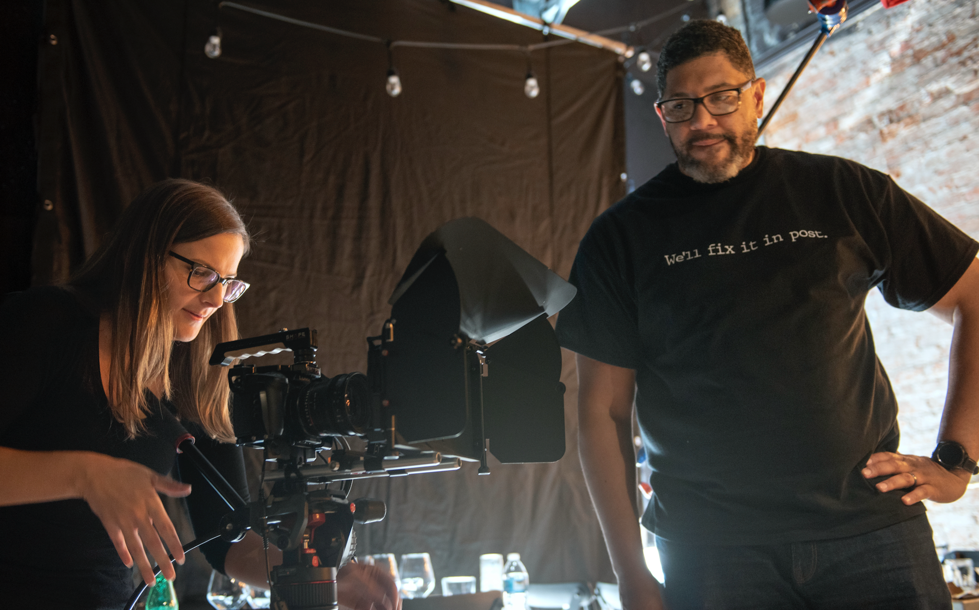 A woman and a man are in a dimly lit room with a black backdrop, working with a professional camera on a tripod; the woman is adjusting the camera, while the man stands nearby with his hand on his hip, wearing a black t-shirt that says 'We'll fix it in post.'