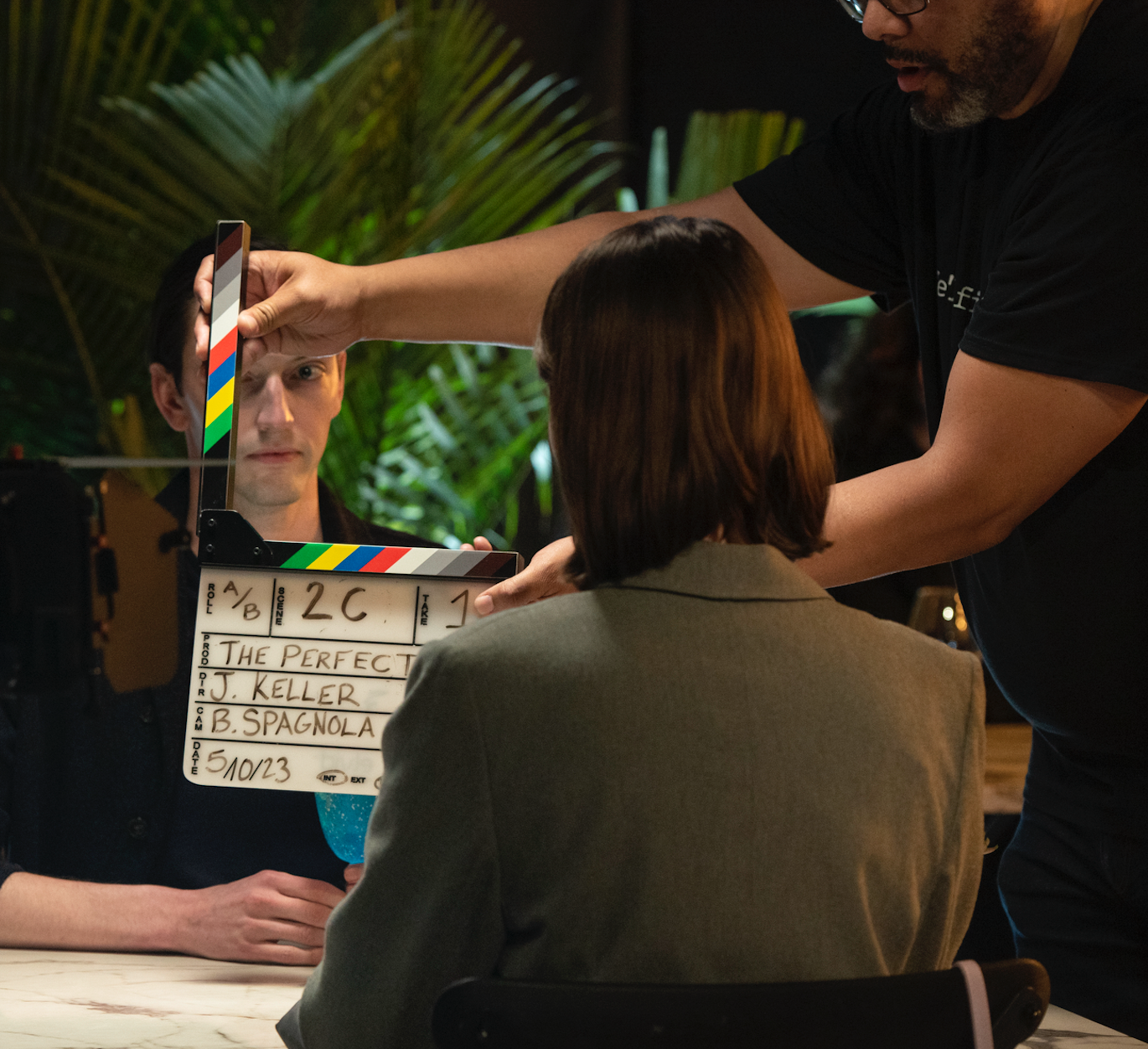 Behind the scenes of a film shoot, a crew member adjusts a clapboard for a scene titled 'The Perfect,' featuring actors J. Keller and B. Spagnola, with a woman in a gray blazer sitting at a table.