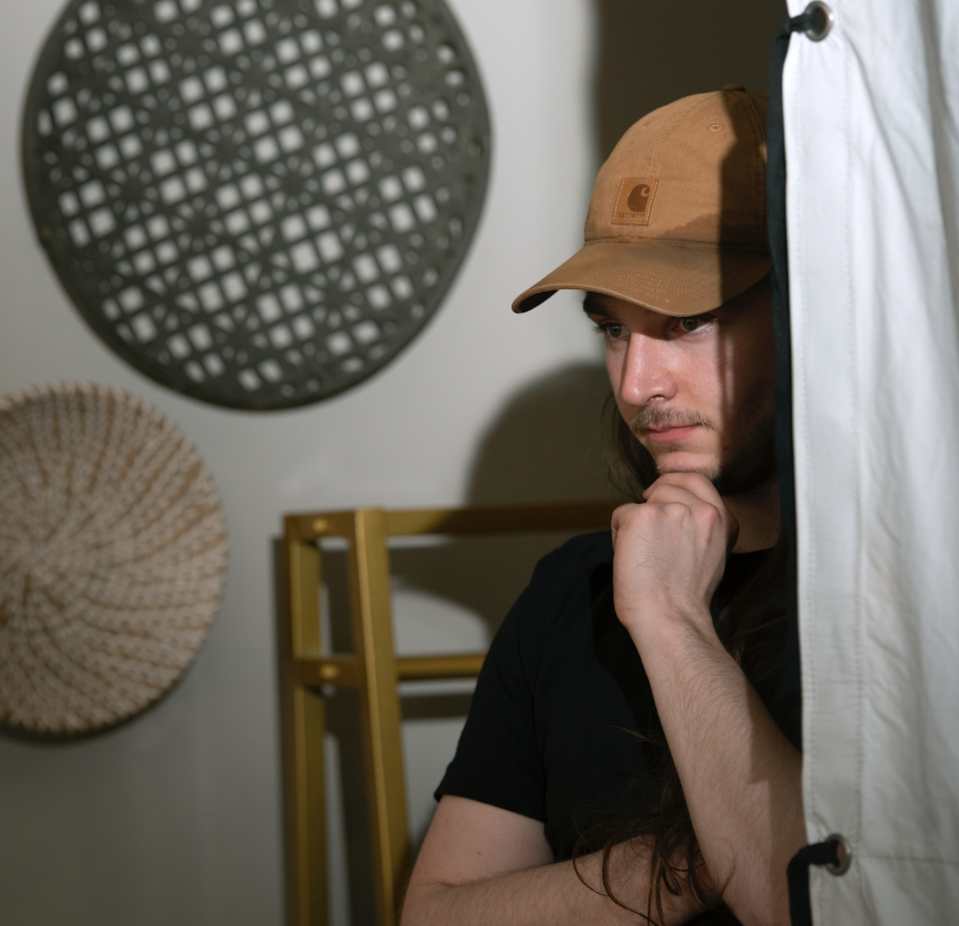 A man in a brown Carhartt cap resting his chin on his hand, appearing thoughtful, standing near a white fabric curtain, with decorative woven wall hangings and a yellow ladder in the background.