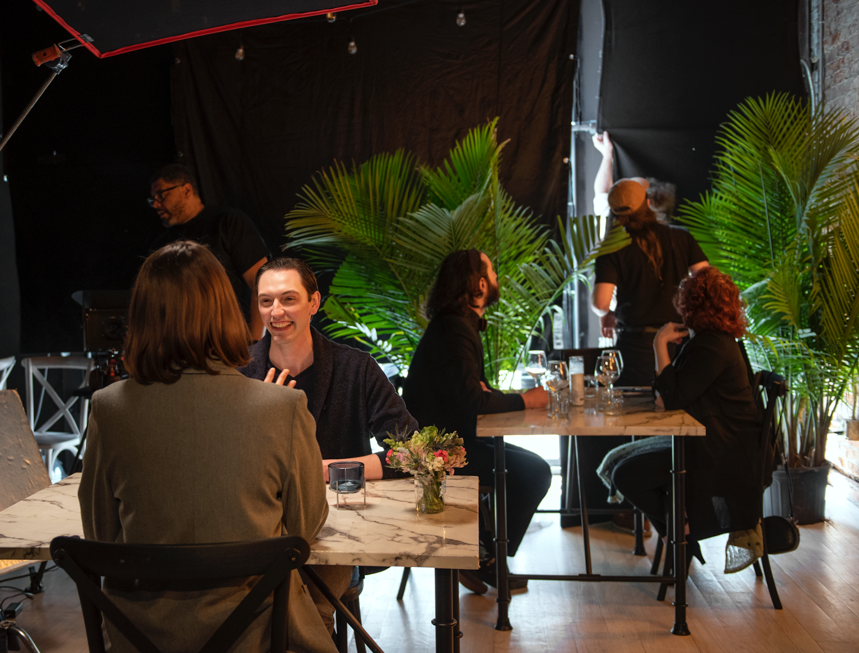 People sitting at tables in a restaurant or cafe, conversing and smiling, with a person in the background adjusting lighting or equipment, and plants as decor.