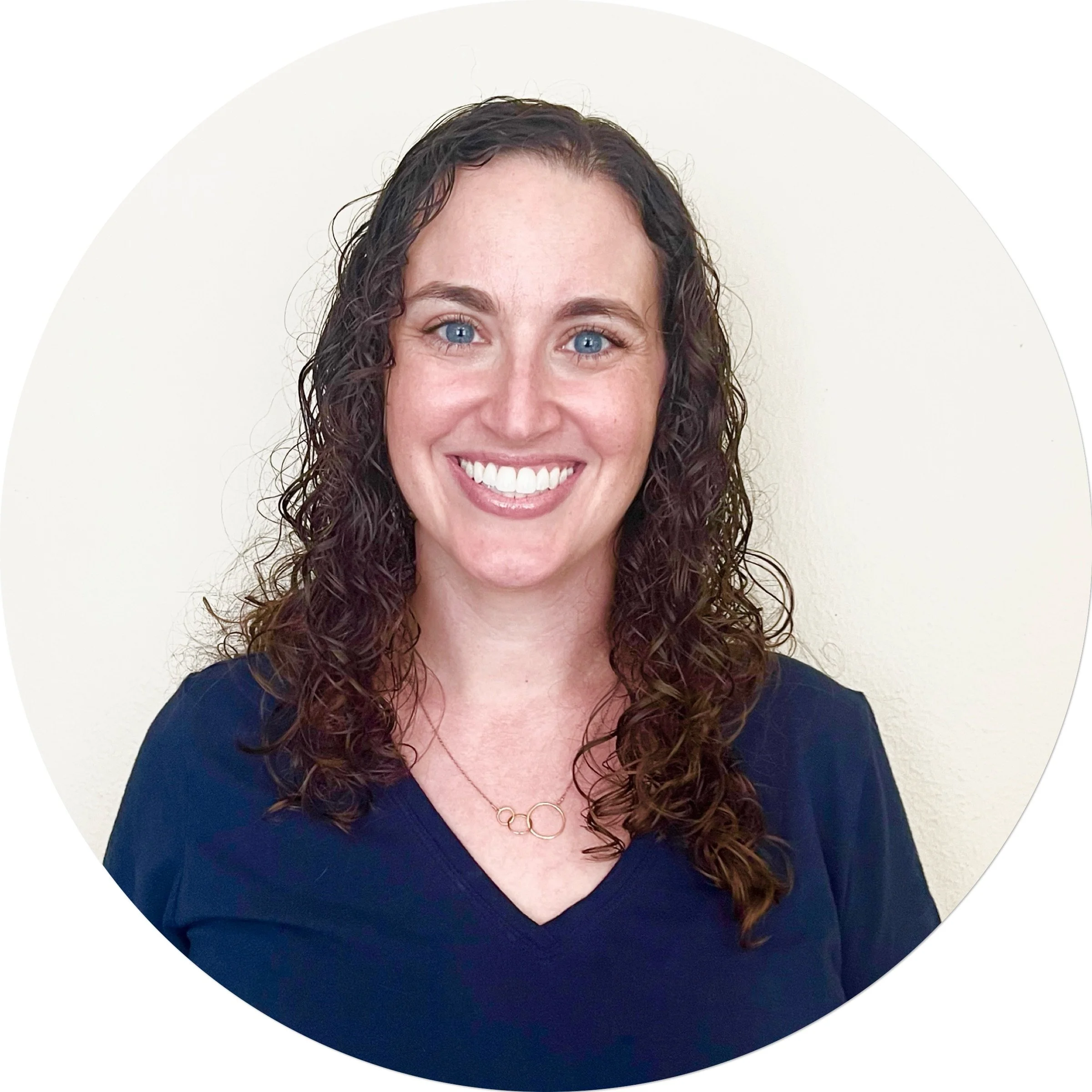 A woman with long, curly brown hair, blue eyes, and a broad smile wearing a navy blue top and a delicate gold necklace, standing against a plain light-colored wall.