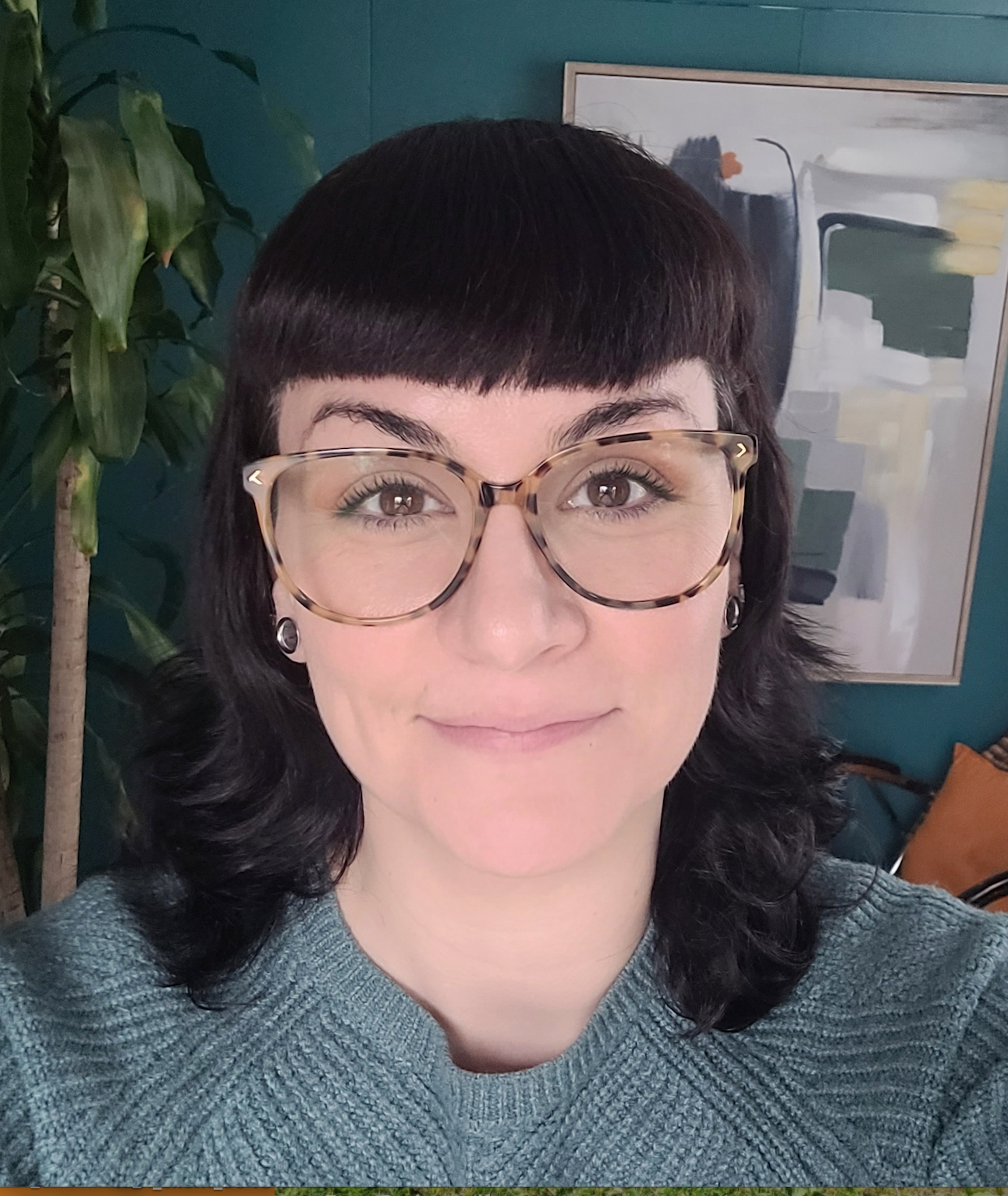 A woman with short dark hair, wearing large tortoiseshell glasses, smiling in front of an abstract painting on a teal wall, with a green plant and an orange cushion visible in the background.