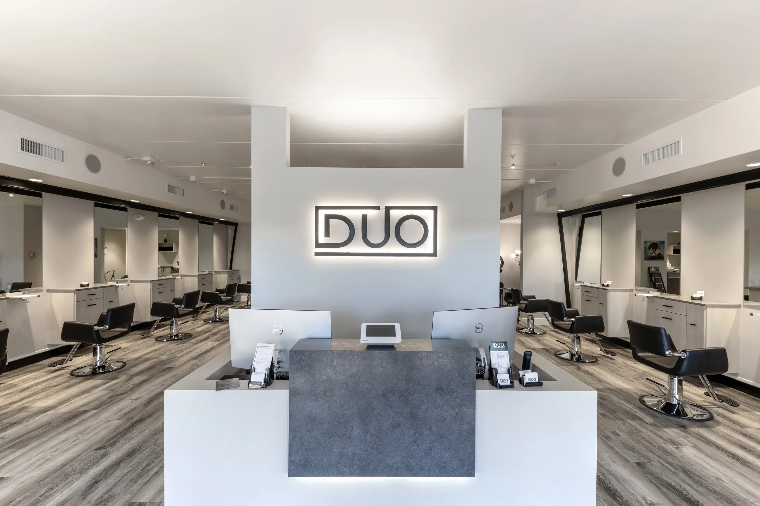 Modern hair salon interior with a sign reading 'DUO' on the wall, black salon chairs, mirrors, and a reception desk in the foreground.