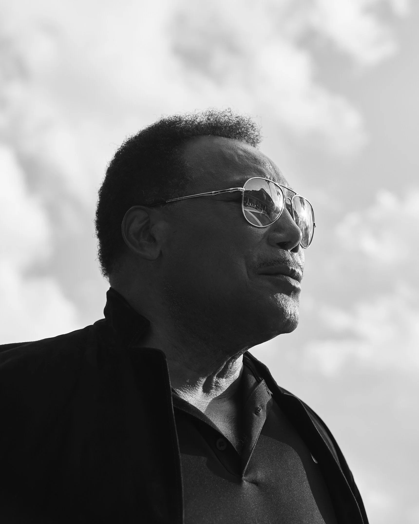 Black and white portrait of a man wearing sunglasses and a collared shirt, looking up against a cloudy sky.