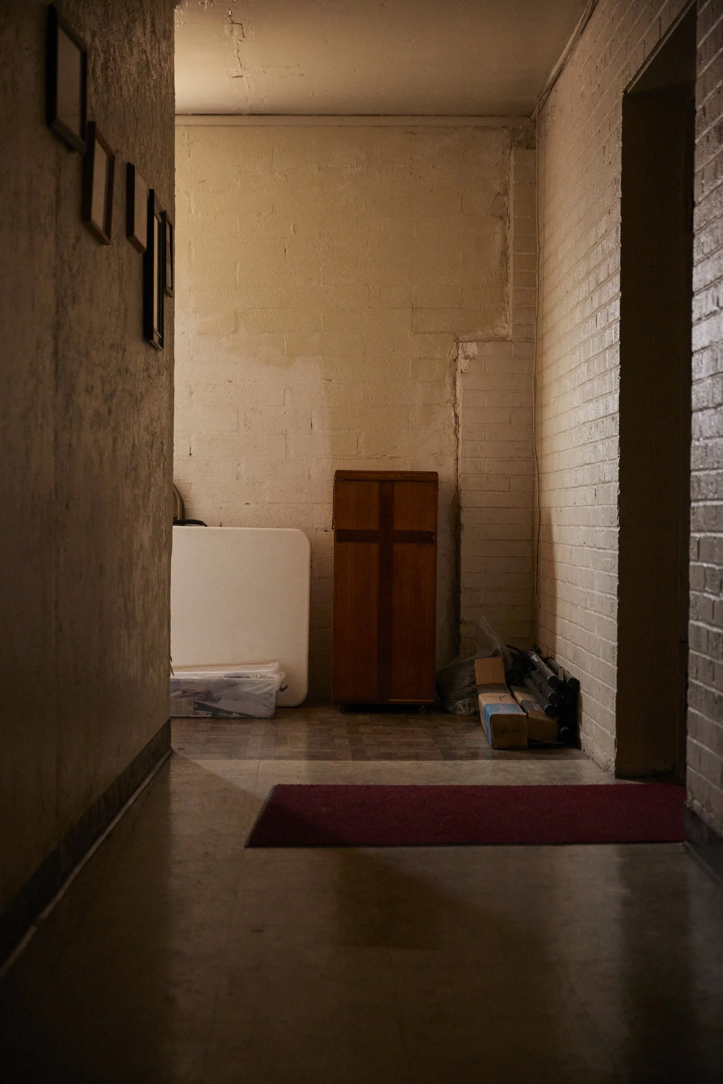 Dimly lit hallway with a wooden cabinet, folded white table, plastic container, and boxes by a brick wall.