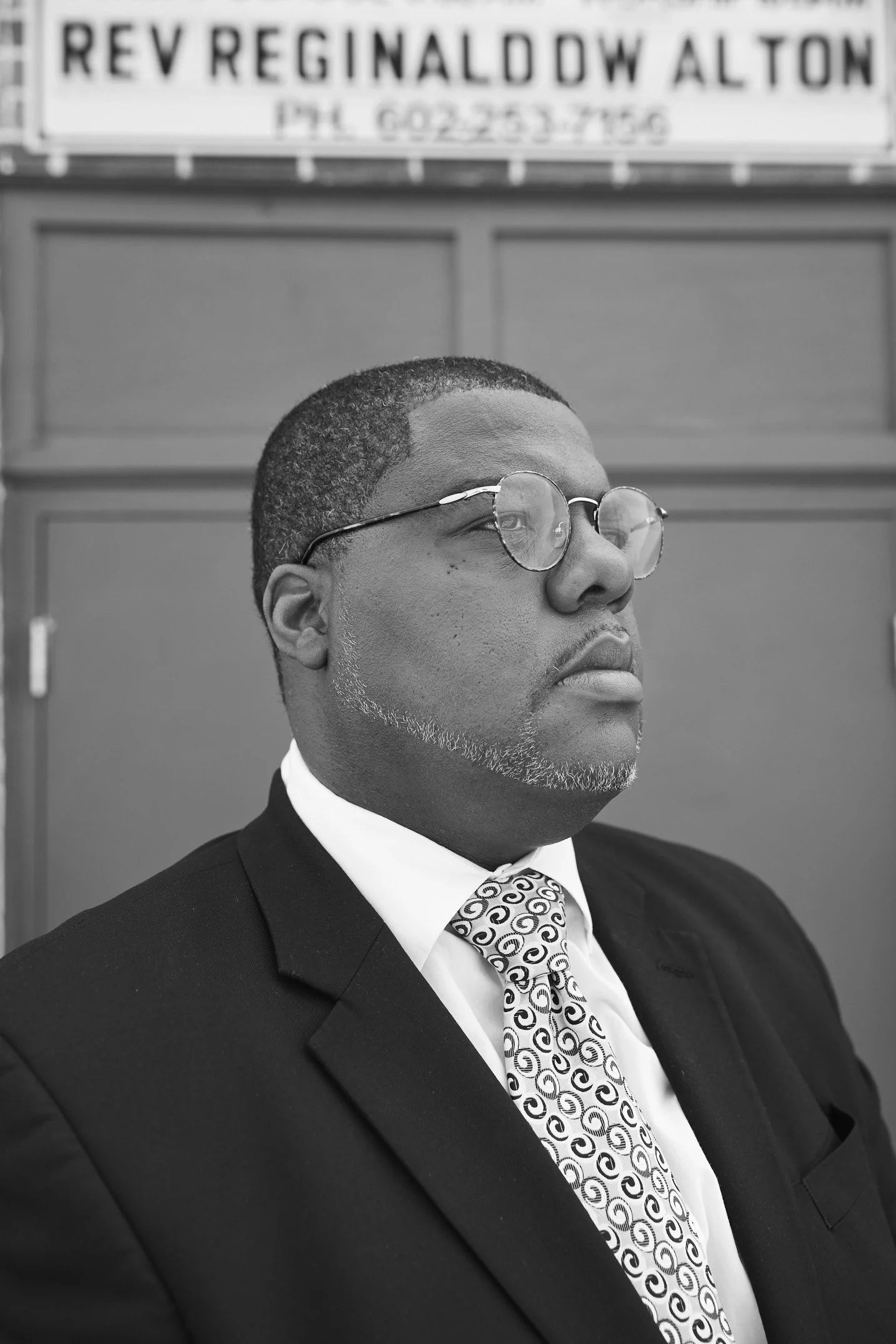 Man in suit with patterned tie and glasses standing in front of a sign reading 'REV REGINALD DW ALTON'