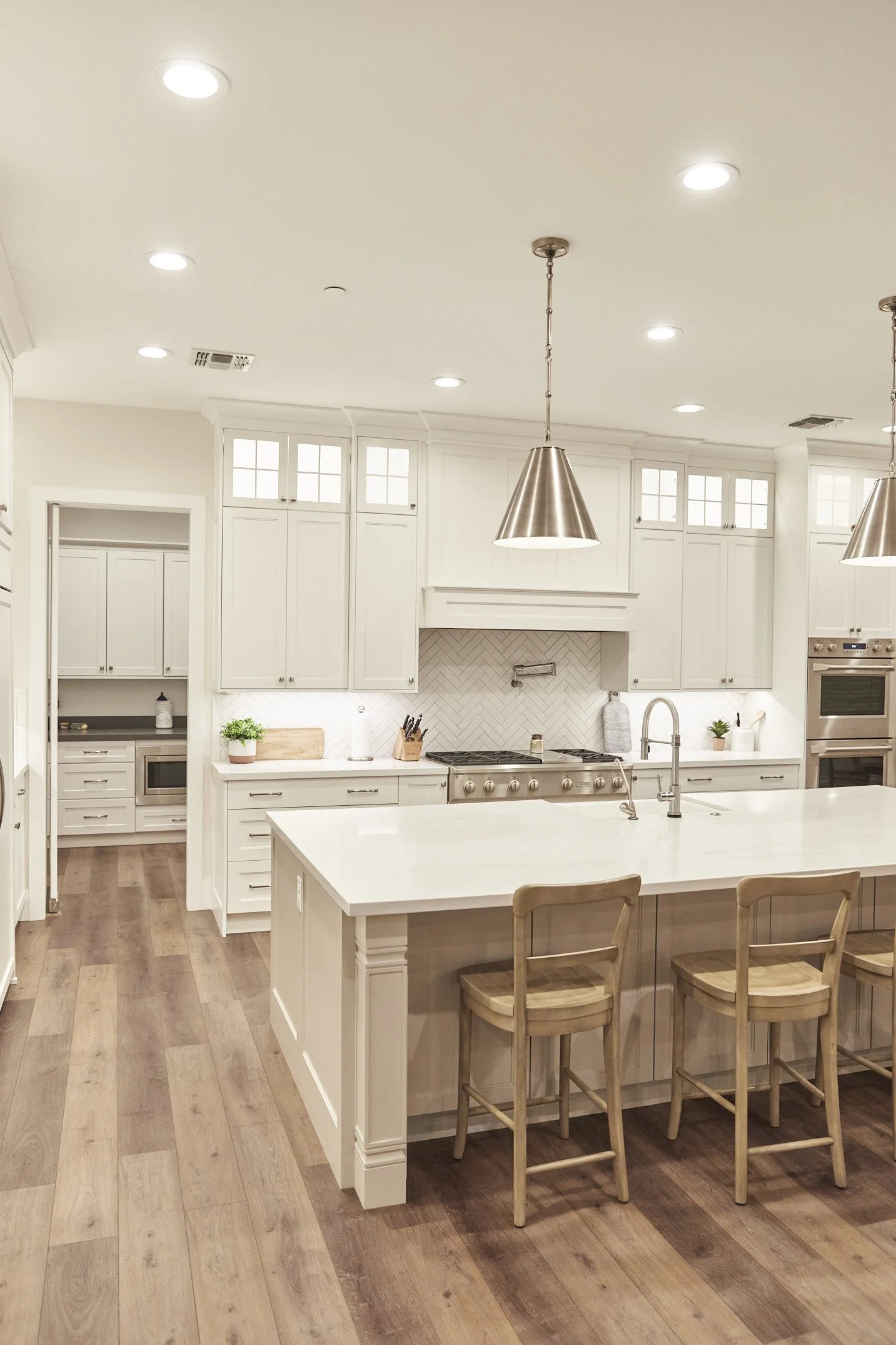 Modern kitchen with white cabinetry, a large island, wooden stools, stainless steel appliances, herringbone tile backsplash, and pendant lighting.