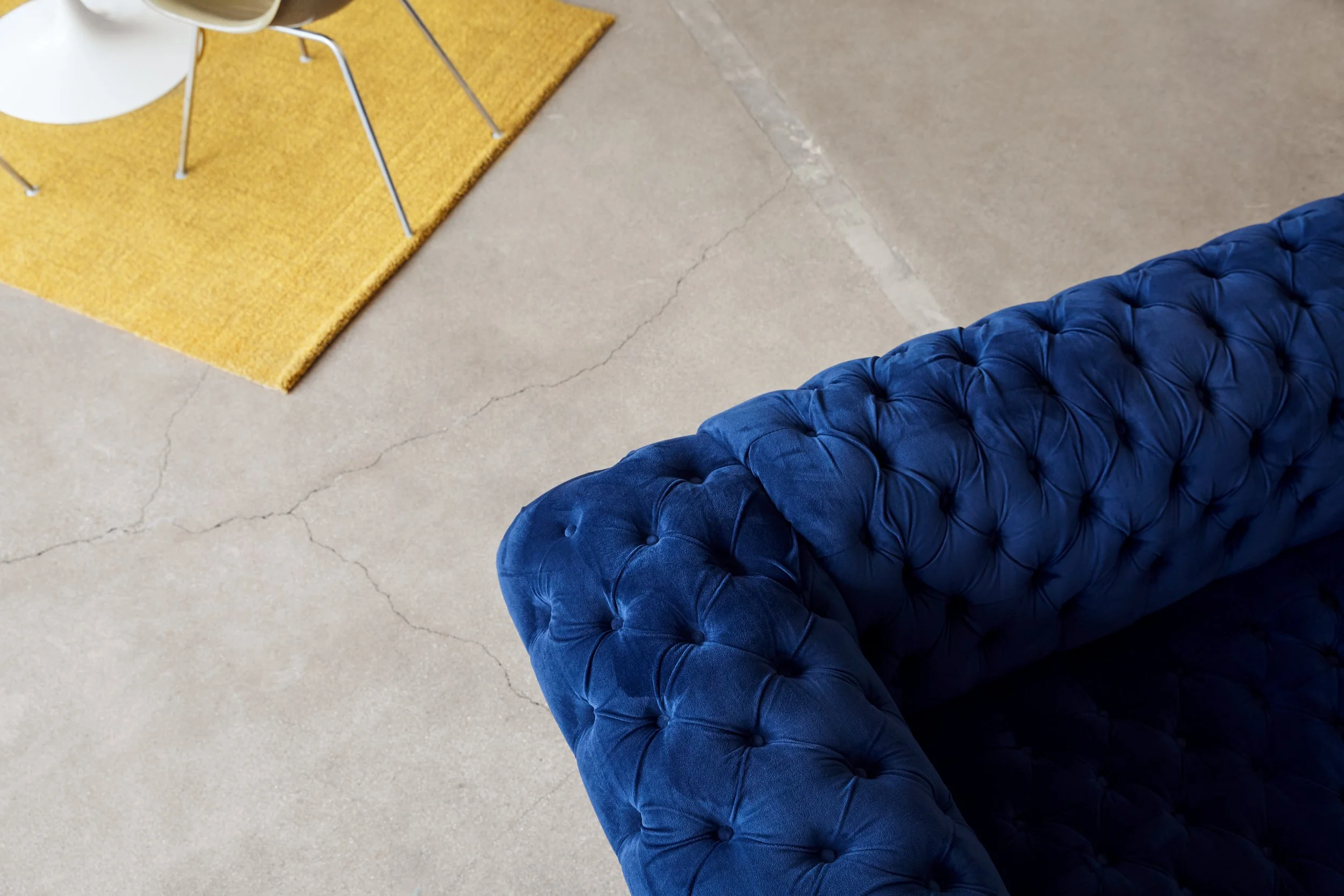 Overhead view of a blue tufted couch, yellow rug, white round table, and chairs on a concrete floor.
