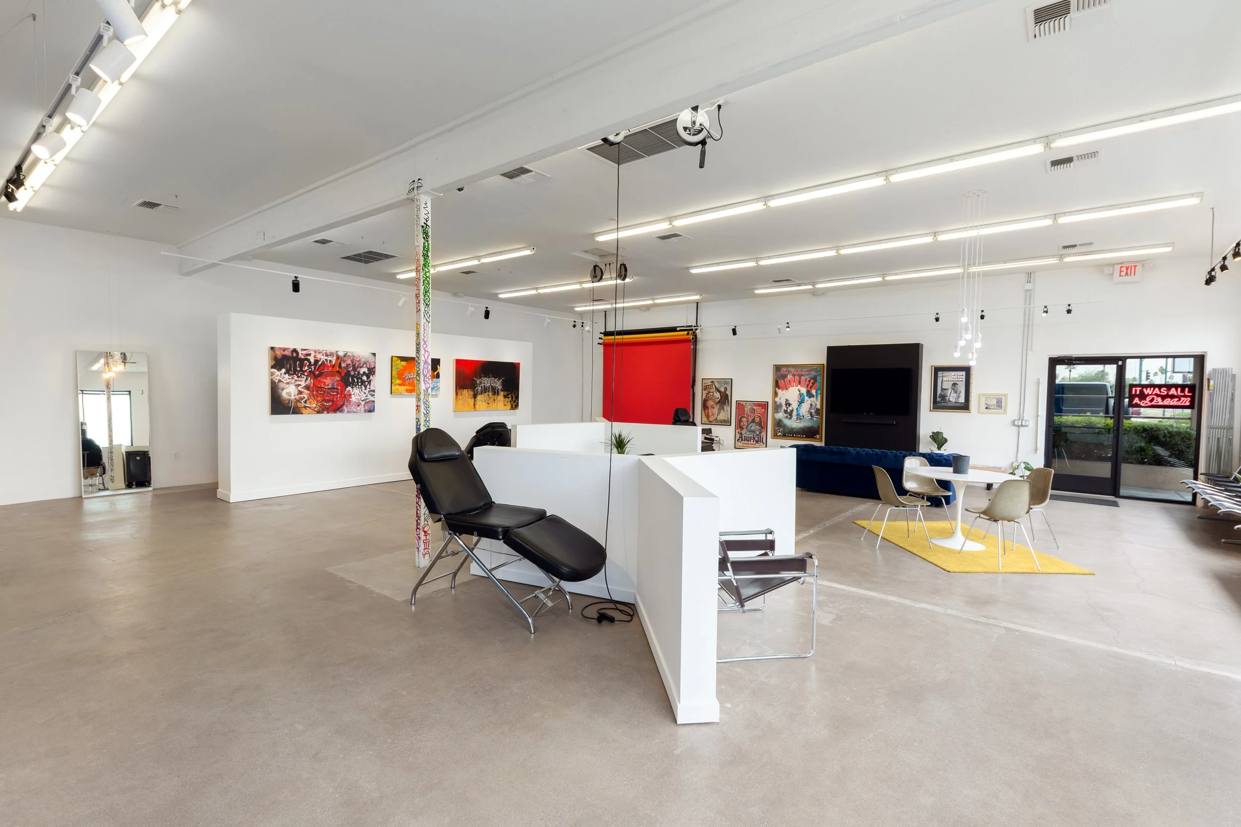 Modern art studio interior with artwork on walls and minimalist furniture, including a black reclining chair, glass tables, and a white circular table with chairs on a yellow rug. There is a large television, studio lights, and a red backdrop. A glass door with a neon sign viewed from outside reads 'IT WAS ALL A DREAM.' The room has white walls and ceiling, with exposed beams and concrete floors.