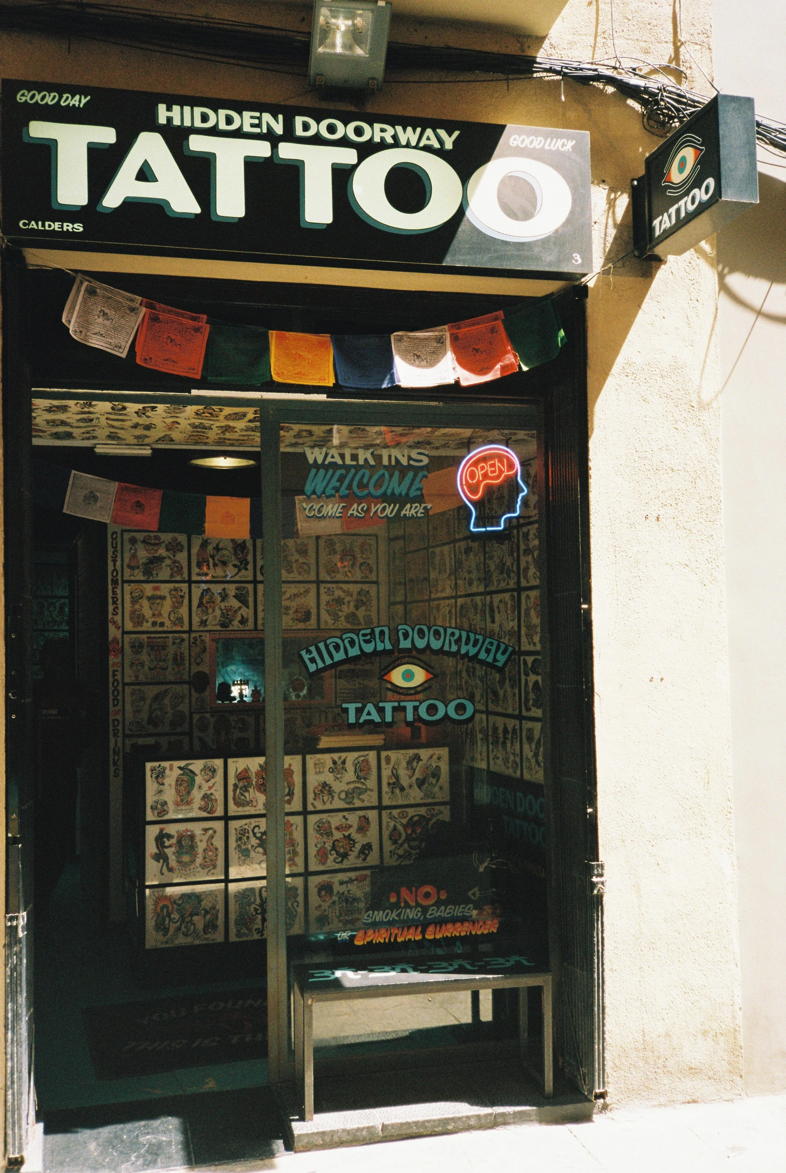 Cartel de una tienda de tatuajes llamada 'Hidden Doorway Tattoo'. Tiene colores negros, verdes y blancos, con un símbolo de un ojo en el cartel y en la placa. También hay un letrero que dice 'Somos bienvenidos a entrar como seas' y luces de neón que dicen 'Open'. La tienda tiene decoraciones tradicionales de tatuajes en la ventana.