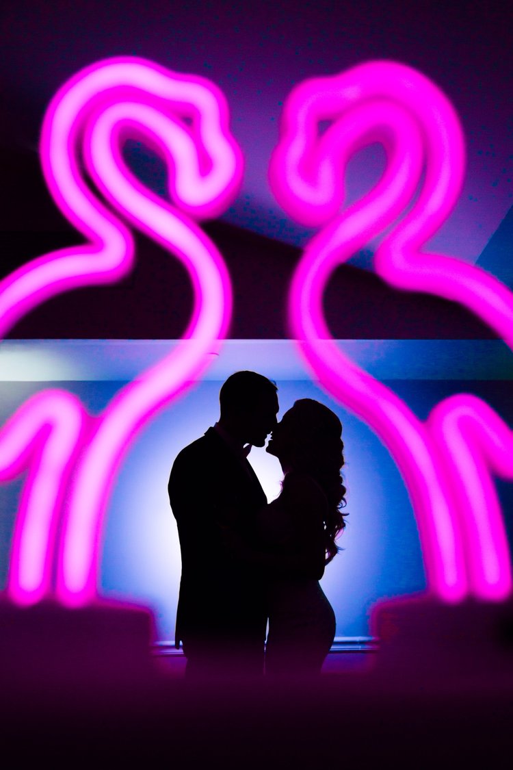 Silhouette of a couple kissing in front of neon pink flamingo signs.