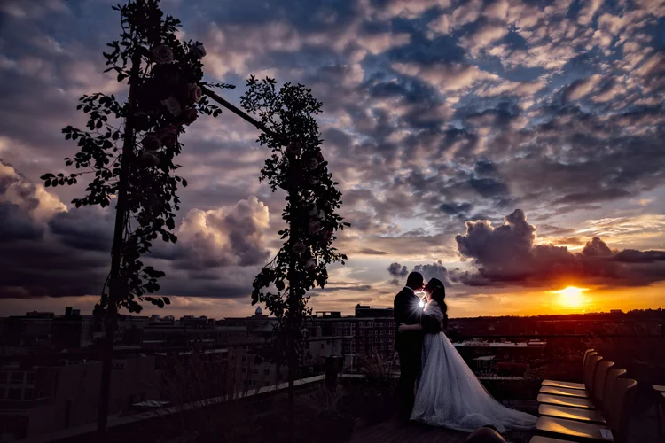Couple in wedding attire sharing a kiss on a rooftop at sunset, with a decorative floral arch and cityscape in the background.